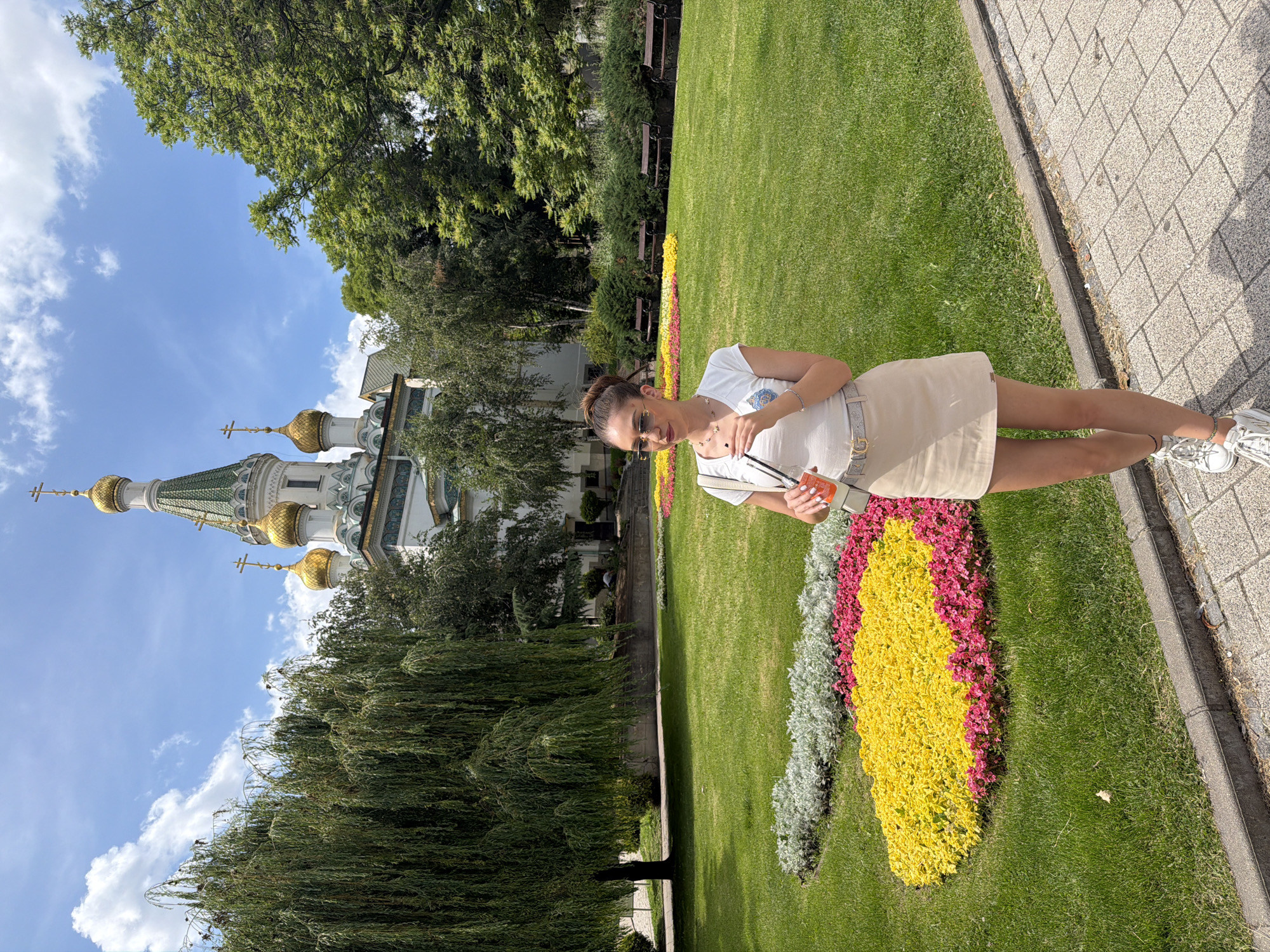 A young woman stands in a park in front of the Alexander Nevsky Cathedral in Sofia, Bulgaria. She is wearing a white t-shirt, a beige skirt, and white sneakers. She has sunglasses on and is holding a phone and a drink. The park has green grass, flower beds with yellow and pink flowers, and trees. Several benches are visible in the background. The cathedral has gold domes and a green roof. The sky is blue with white clouds.