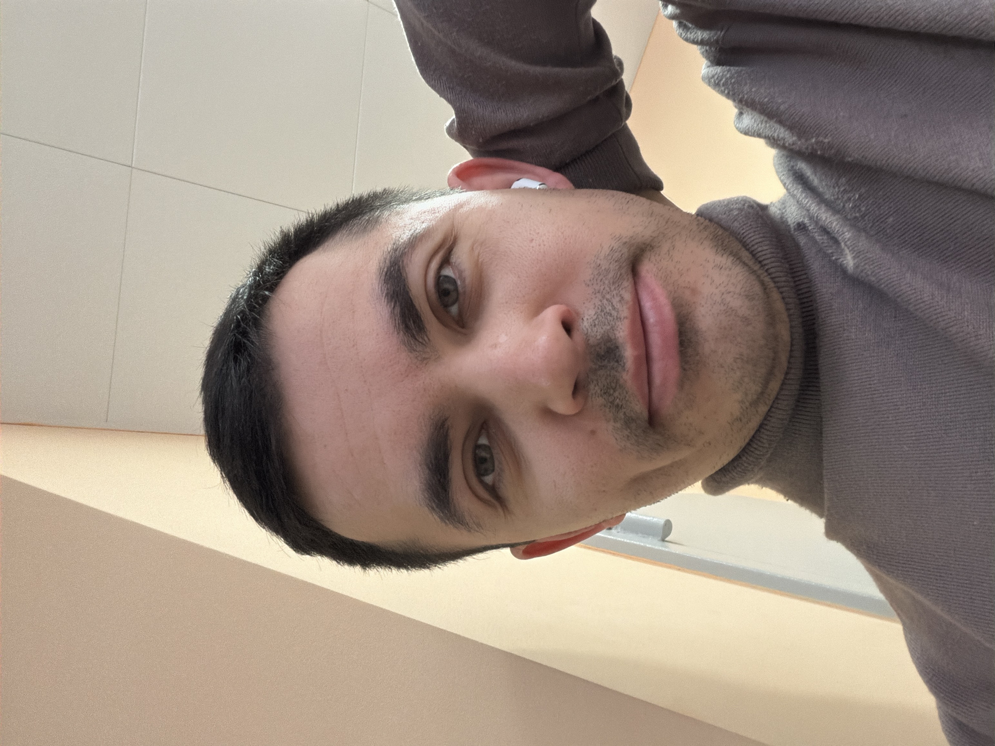 A close-up selfie captures a man with short dark hair, light eyes, and stubble. He wears a grey turtleneck sweater. A white wireless earbud is visible in his left ear. The background consists of tiled walls and a painted shelf, suggesting an indoor location. The image provides no specific information to indicate the location is Izhevsk, Russia.