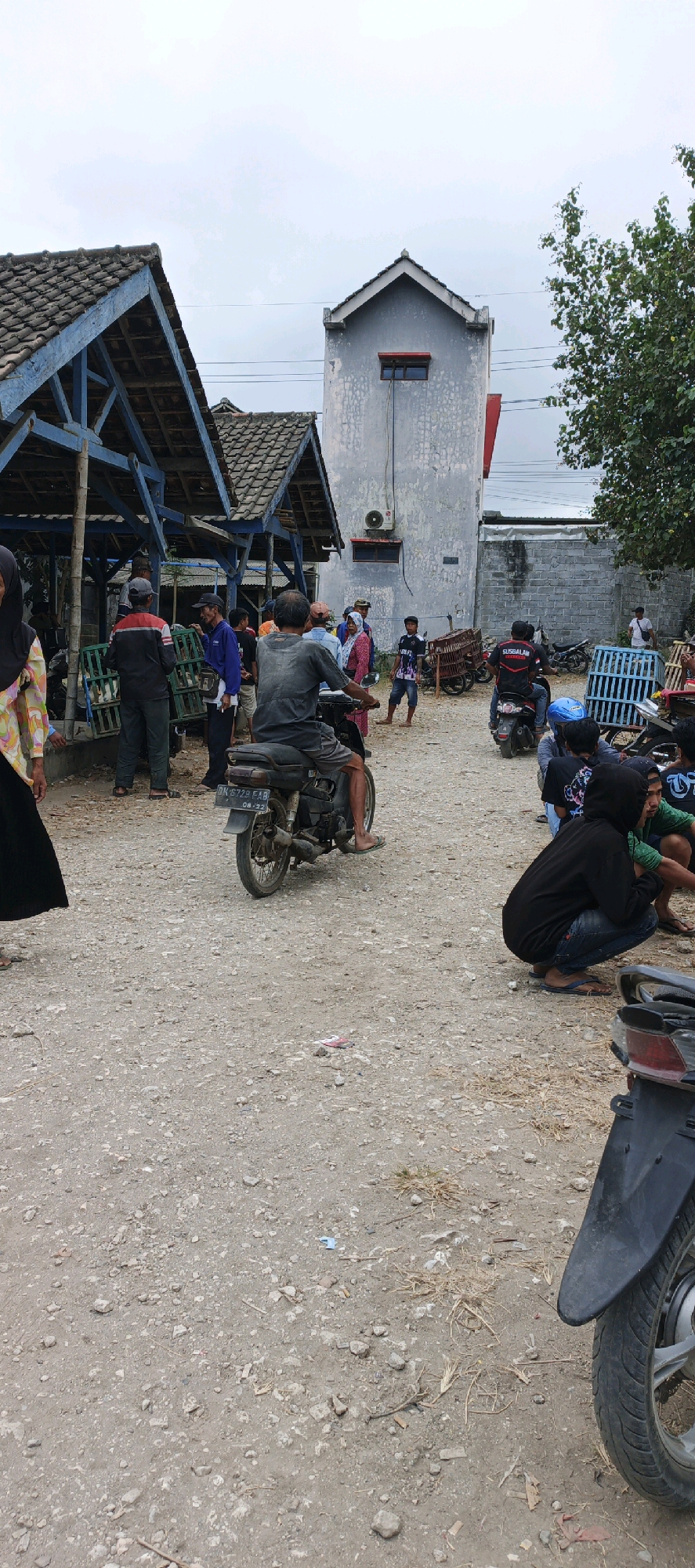 The image captures a scene in Kasiyan, Indonesia, likely a market or a gathering area. In the foreground, a person is seated on a motorcycle, facing away from the camera, with their legs visible. Another motorcycle is parked in the lower right corner, its rear wheel and fender prominent. The ground is a rough, gravelly surface.
In the mid-ground, several people are gathered. A man in a grey t-shirt and shorts is riding a motorcycle towards the right. To the left, a group of people are standing near a blue-roofed structure with a tiled roof, which appears to be a stall or shelter. Some individuals are observing or interacting with items within this area, possibly merchandise. A man in a black jacket with red accents is seen talking to another person. Further into the scene, a tall, narrow building with a grey facade and a red-topped roof stands, with a visible air conditioning unit on its exterior. More motorcycles are parked in the background, and several individuals are visible in various states of interaction and observation, including a person in a black hoodie squatting on the ground. The overall environment suggests an outdoor public space with a mix of residential and commercial elements, under an overcast sky.