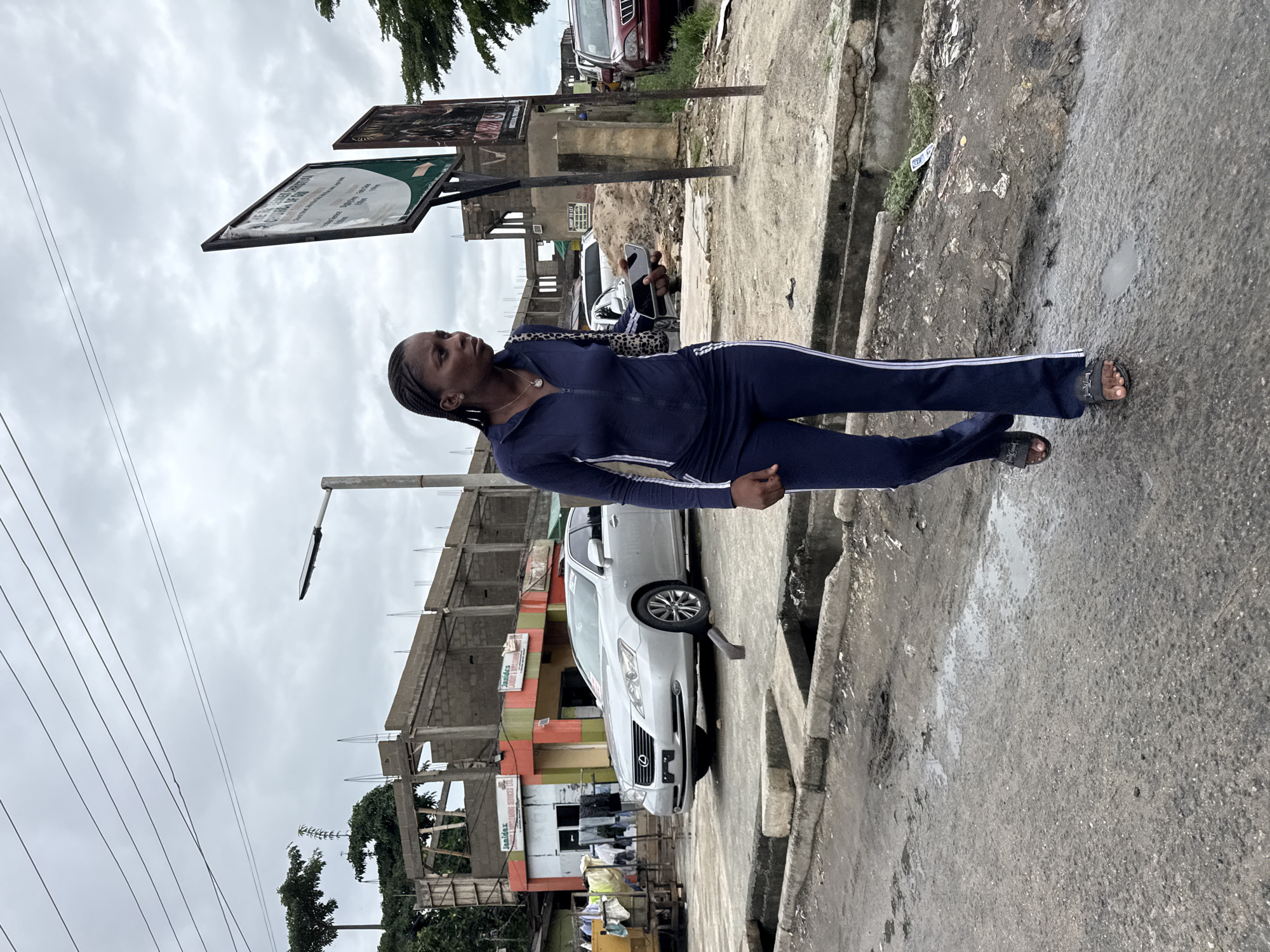 A young woman stands on a concrete street in Ado, Nigeria. She is wearing a dark blue tracksuit with white stripes down the sides. Behind her is a white car, and behind the car is a building under construction with some finished commercial space on the ground floor. In the background, there is a signpost. The sky is overcast. The ground is wet.