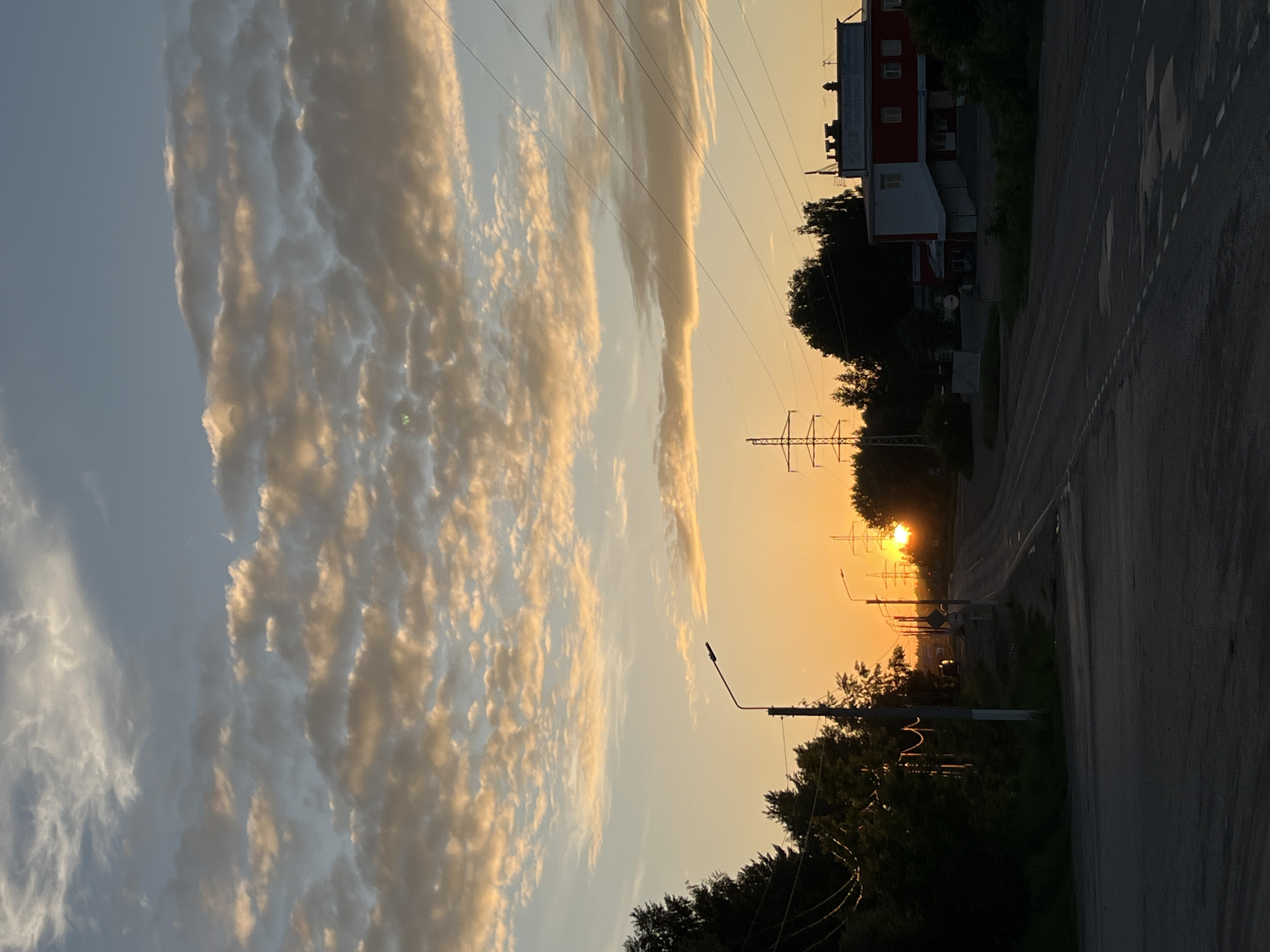 The image captures an urban landscape in the city of Chaikovsky, Russia, during what appears to be sunset, characterized by low, warm light. The upper two-thirds of the frame is occupied by a sky dominated by altocumulus clouds, illuminated from below in shades of gold, orange, and light brown, contrasting with areas of light blue and grey. The sun, a bright orange disc, is visible low on the horizon in the lower-mid right section of the image, partially obscured by dark foliage.
In the foreground, a dark asphalt road, likely a two-lane thoroughfare, recedes into the distance. White dashed lane markings are visible on its surface, along with several dark, irregular patches, possibly indicating standing water or repairs.
The midground features a dense line of dark green trees and foliage running along both sides of the road. On the left, a tall, dark streetlamp with a curved arm and a single luminaire extends over the road. Several dark utility poles are scattered along the roadside, supporting multiple power lines that traverse the sky horizontally and diagonally.
On the right side of the road, a two-story building is visible, featuring a red upper facade and a white lower section, with multiple rectangular windows. Cyrillic text is discernable on a sign positioned above the red facade. Further into the distance, two prominent metal lattice power transmission towers (pylons) are silhouetted against the bright horizon, one larger on the right and another partially visible on the left. No individuals or actively moving vehicles are present within the frame.