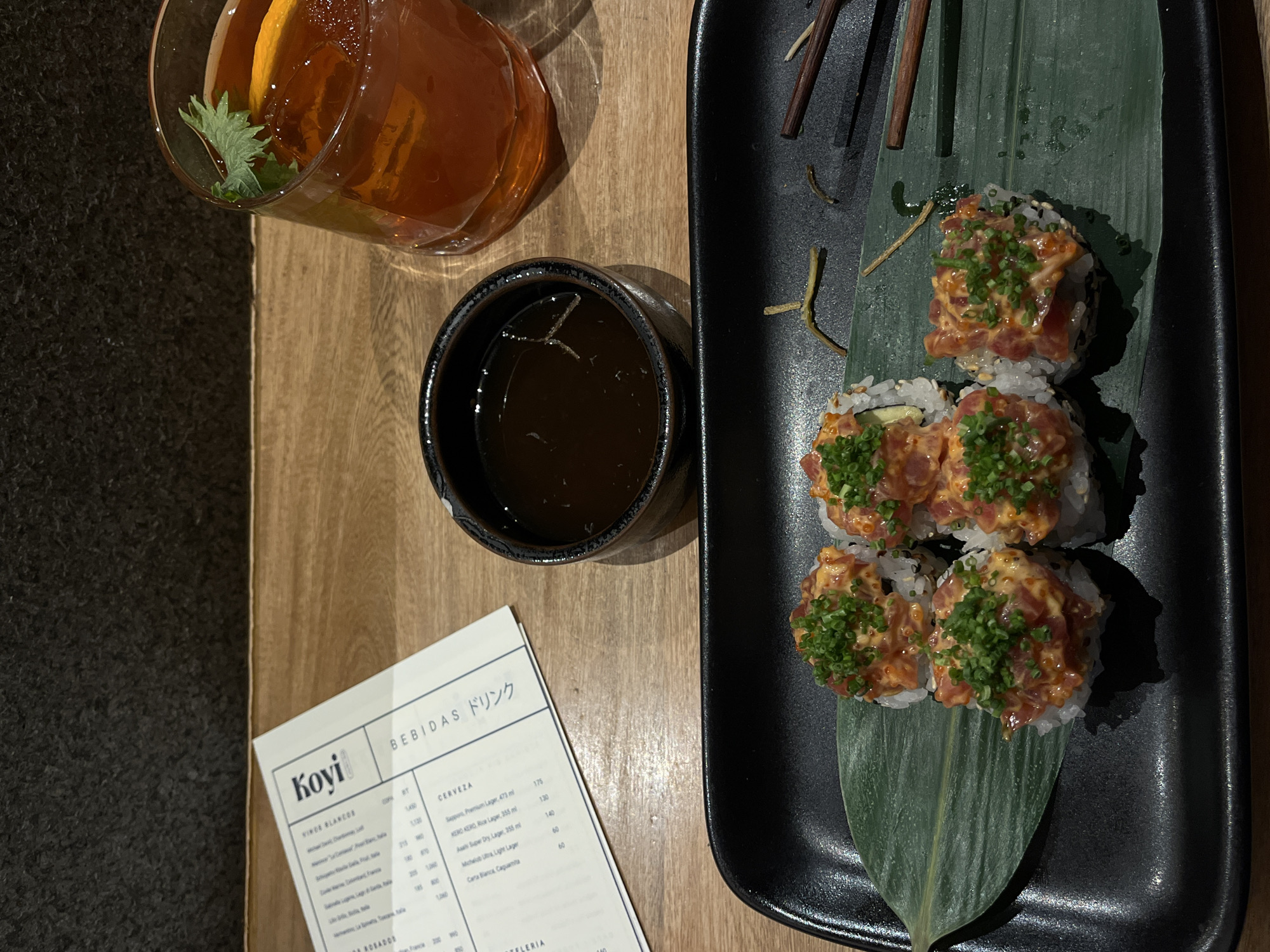 The image shows a restaurant table, likely in Mexico City, Mexico, as indicated by the restaurant name "Koyi" on the menu and the "Bebidas" heading in Spanish and Japanese, a cocktail with an orange slice and a sprig of mint, a small bowl of dark sauce, a black rectangular dish with sushi rolls topped with chopped greens, arranged on a large leaf, and chopsticks resting on the dish. The table is made of wood and the menu is partially visible at the bottom of the frame.