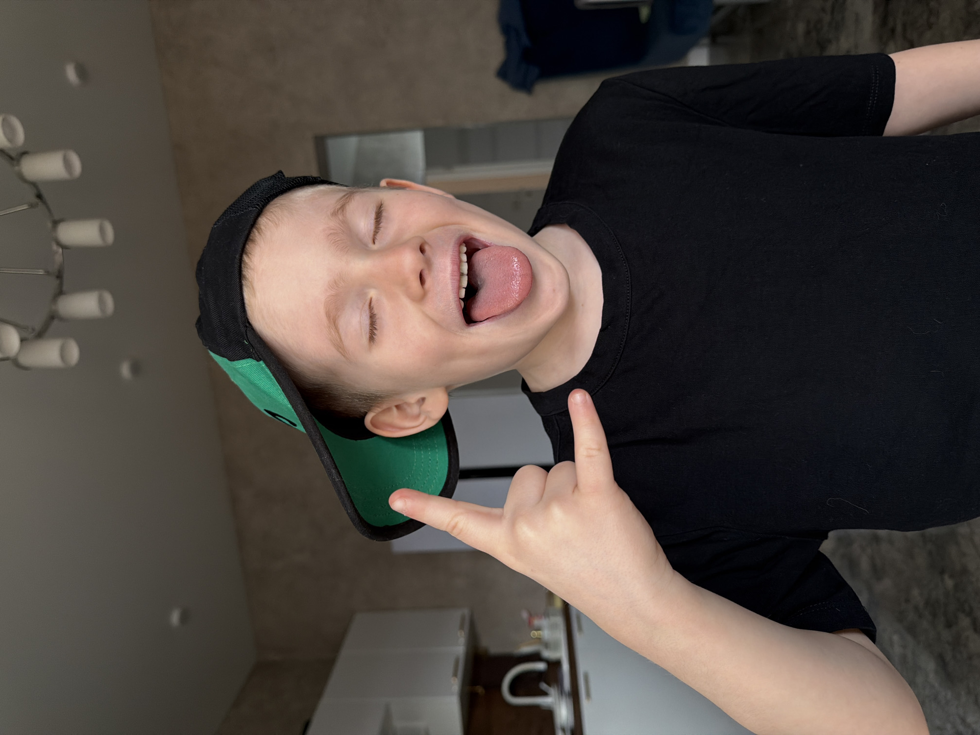 A child with fair skin is facing the camera and sticking out his tongue. He is wearing a black t-shirt and a backwards baseball cap with a black top and a green brim. He is making the "rock on" hand gesture. In the background, there is a chandelier with multiple lights, a kitchen sink, and cabinets. It is most likely taken inside a home in Rossiyskiy, Russia.