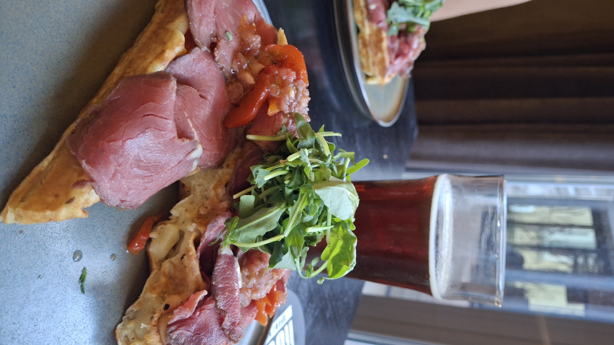 In a close-up, eye-level shot, a waffle sandwich topped with thinly sliced roast beef, diced tomatoes, and a pile of arugula sits on a light blue plate. The waffle is golden brown and cut into wedges. A glass of amber-colored beer, about two-thirds full, stands behind the sandwich. The background is out of focus, with dark brown curtains to the left and a blurry window with vertical panes and outdoor reflections to the right. The setting appears to be an indoor dining area.