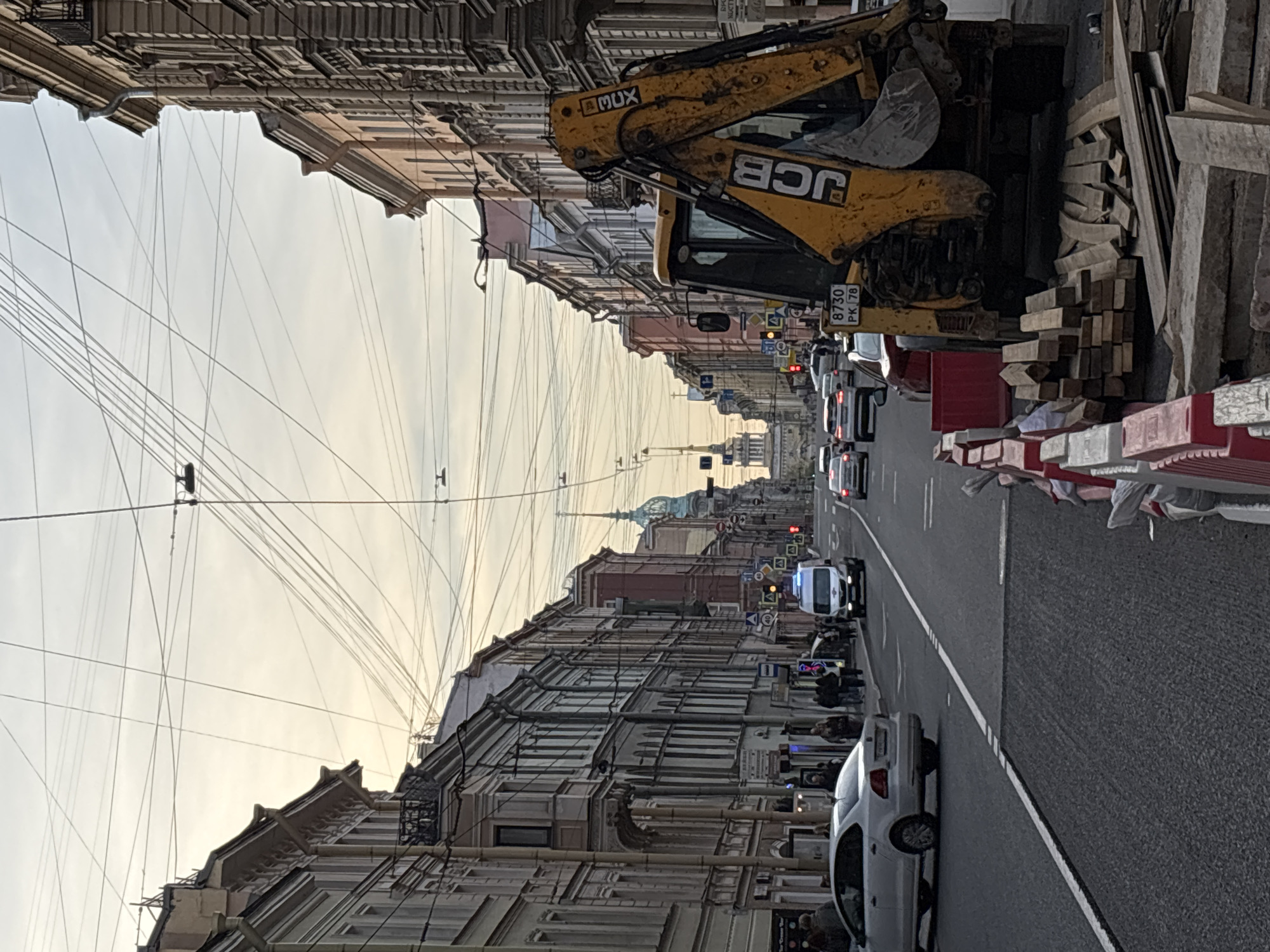 The image shows a street scene in Saint Petersburg, Russia. A yellow JCB excavator is parked on the right side of the street, partially blocking the view. Wooden planks are piled nearby, behind red and white road barriers. A line of vehicles, including a white car, a white van with flashing blue lights, and several trucks, are visible on the road. Pedestrians can be seen walking along the sidewalk. Overhead, numerous electrical wires crisscross the sky. In the distance, a building with a prominent golden spire can be seen. The buildings lining the street are multi-story and have intricate architectural details. The sky is overcast and light. The scene appears to be a construction site or road work in progress.