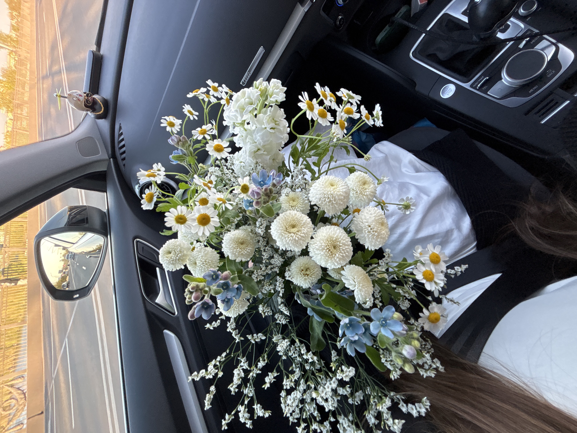 A close-up, high-angle photograph shows a bouquet of flowers resting inside a car. The bouquet features white and yellow daisies, white pompom-like flowers, white stock, and small blue flowers with sprigs of tiny white blossoms. The bouquet is primarily positioned on the dashboard and extending towards the passenger seat, which has a white fabric covering it. A black garment also lies on the seat. Parts of the car interior, including the dashboard, a side mirror reflecting a street scene, and the car's console with dials and buttons, are visible. A small decorative item is attached to the dashboard near the windshield. A strand of brown hair is visible at the bottom of the image. The photograph does not confirm a specific location such as the city of Krasnoyarsk, Russia.