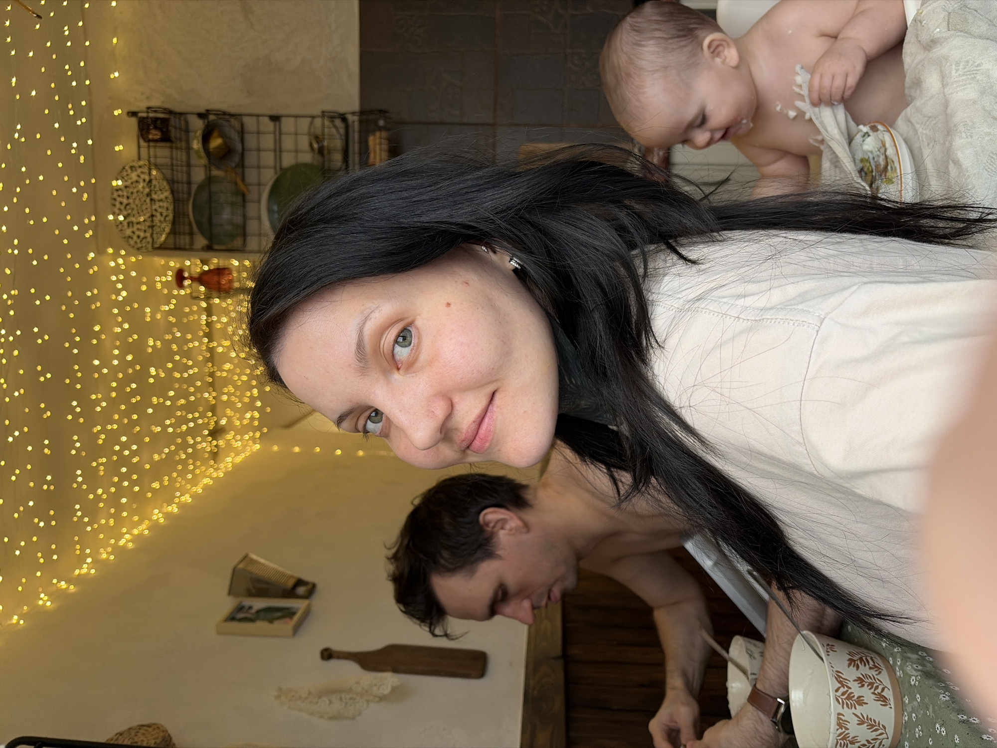 The image depicts a family scene indoors. A woman with dark hair is in the foreground, smiling at the camera. Behind her is a man bending over, and a baby smeared with what appears to be cream is being held. The environment includes a wall decorated with string lights and kitchenware. The presence of wooden decor elements and kitchen utensils suggests the scene is set in a home environment in the city of Chaikovsky, Russia. The baby is being cleaned with a cloth by its parents, indicating a family event or mealtime.