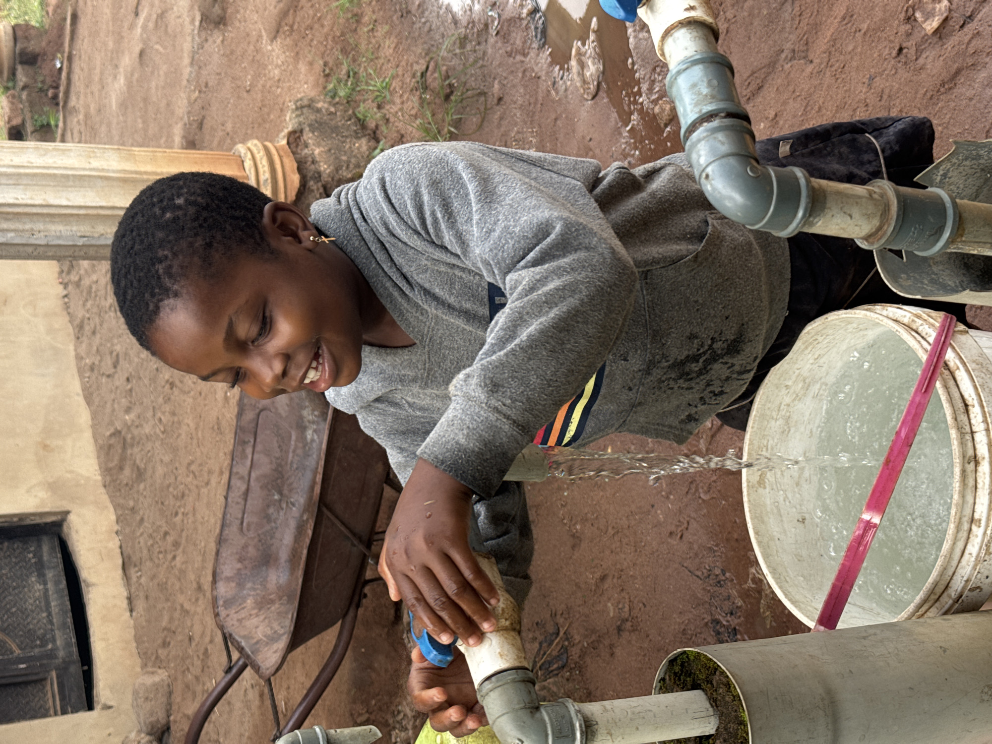 The image captures a smiling young person, likely a child or teenager, collecting water from an outdoor faucet. The subject, with short dark hair and a gold cross earring in their left ear, wears a grey hooded sweatshirt with a prominent colorful striped pattern across the chest. Their hands are clasped around the white and blue faucet, from which a steady stream of clear water gushes into a large, white plastic bucket positioned directly below. The bucket is already partly filled, and a red stick or handle is visible inside it.
The scene takes place outdoors on unpaved, reddish-brown earthen ground, which appears damp and slightly muddy in places due to splashing water. Patches of green grass are visible in the background. The water source consists of grey and white PVC pipes, some connected with blue fittings, forming an exposed plumbing system. One pipe near the ground shows some moss or algae growth.
In the background, a rustic building with light-colored walls and a dark, shuttered window or doorway can be seen. A rusty brown wheelbarrow is also partially visible on the left side of the frame. The overall lighting is bright and even, indicating daytime. The person's wide smile conveys happiness or contentment while performing the task. No discernible text is visible in the image. The setting suggests a common water collection point in a community like Eziudo, Nigeria.