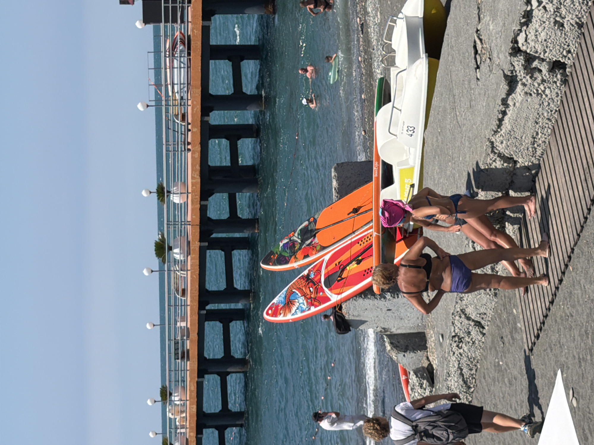 The image depicts a sunny day at a beach in Sochi, Russia, likely during the summer tourist season. In the foreground, several people are present on a dark-colored beach area with some wooden decking. To the left, a young male with a backpack is walking away from the camera. In the center, two women are standing and another woman is seated. Both standing women are wearing bikinis, and the seated woman is also in swimwear.
Dominating the middle ground are two vibrantly decorated paddleboards, one red and one orange, leaning against a concrete structure. Behind them, the azure sea is visible with several people swimming and playing in the water. A small, inflatable ring is also present in the water.
In the background, a pier extends into the sea, supported by dark pillars. The pier is lined with railings, planters containing small green trees, and streetlights. The overall atmosphere suggests a typical day at a seaside resort.