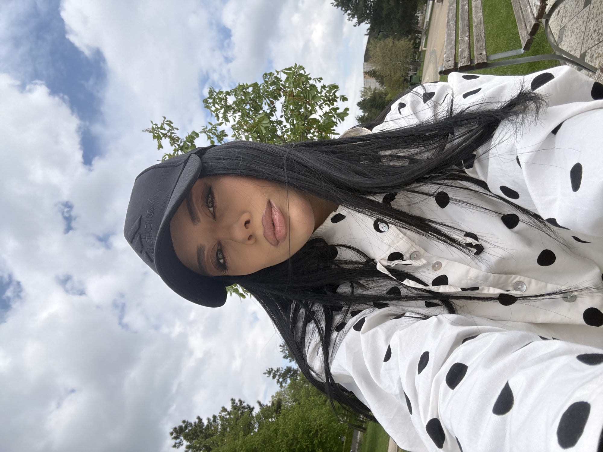 A woman is photographed outdoors in daylight against a backdrop of trees, grass, and cloudy skies. She wears a black cap, and her black hair falls over a white collared shirt with black polka dots. She is angled to the left, with her gaze directed at the camera. The location appears to be a park or green space.
