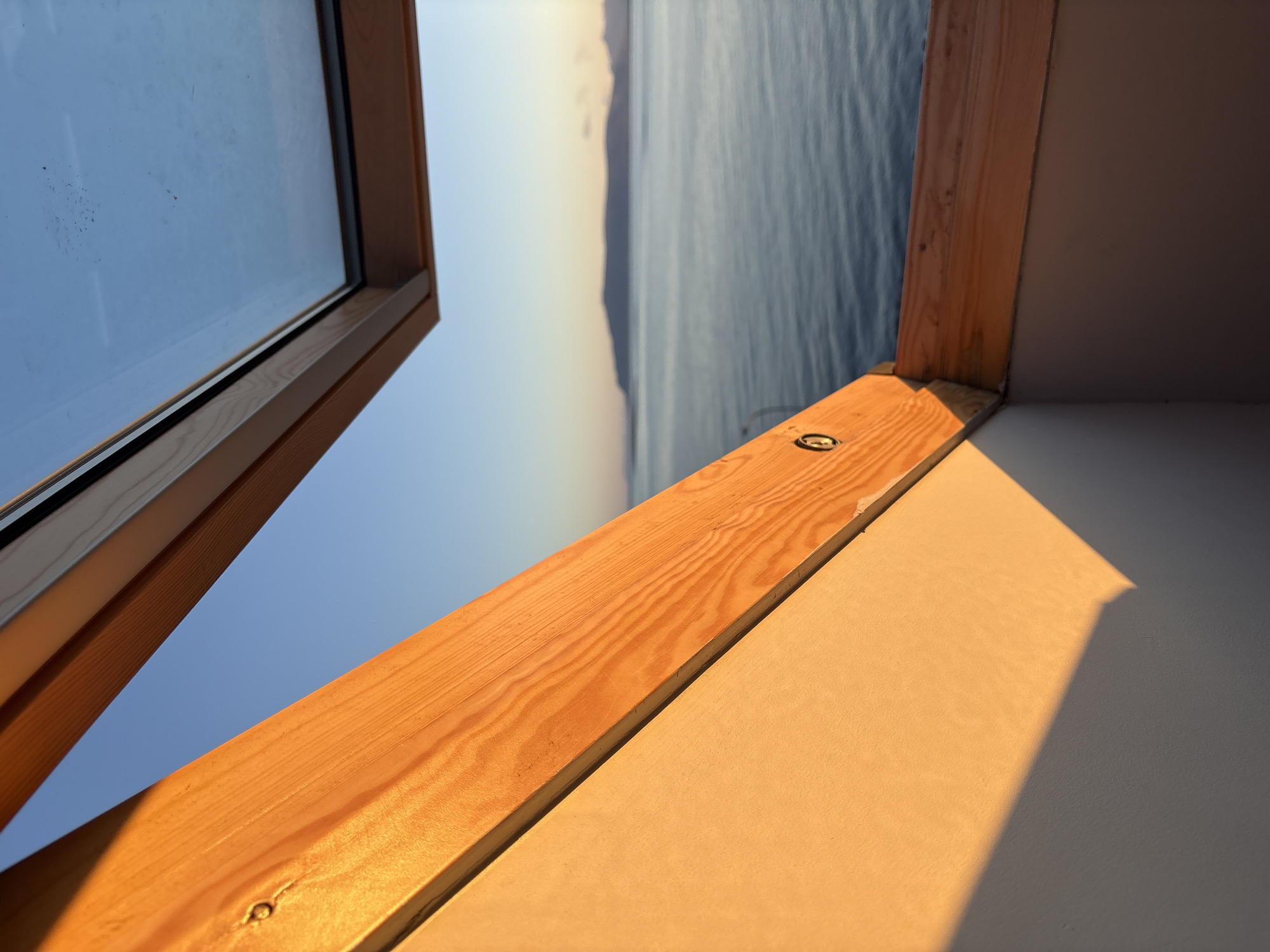 The image presents a view through an open skylight, framing a serene seascape with a distant coastline under a clear blue sky. The foreground is dominated by the warm, golden light of what appears to be either sunrise or sunset, illuminating the angled wooden beams and the textured surface of a wall. The wood grain is prominent, showing natural patterns and a few visible screw heads. A sharp shadow casts diagonally across the illuminated wall. Beyond the window frame, the calm, dark blue water stretches towards a low-lying landmass, its form softened by the distance and atmospheric haze. The overall impression is one of tranquility and natural beauty, with the architectural elements of the skylight providing a contrast between indoor space and the expansive outdoor vista. The specific location within Reykjavik, Iceland is not discernible from the visual information provided in the image itself.
