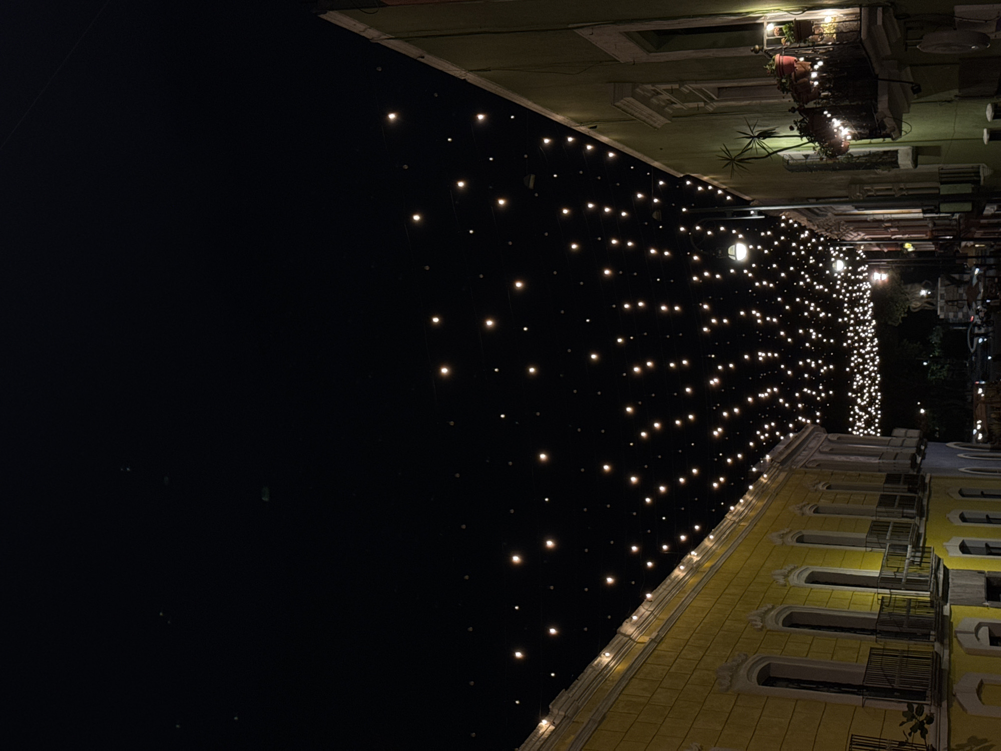 At night, the camera faces an ornately designed yellow building in Puebla, Mexico. Strings of lights are draped in front of the building. Across the street, other buildings decorated with lights can be seen. The lights are most dense on the street facing the camera.