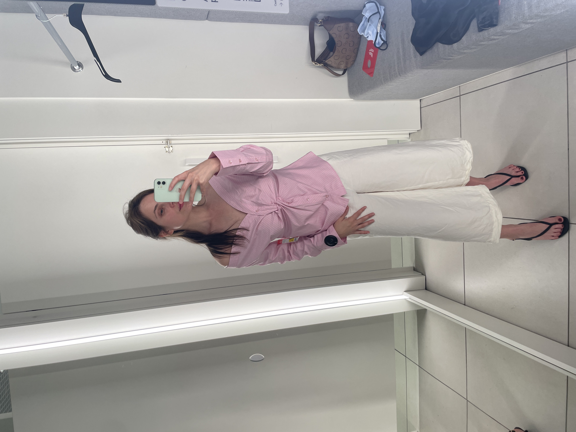 A woman is standing in a fitting room, taking a mirror selfie with her phone. She is wearing a pink and white striped off-the-shoulder shirt with a knot in the front, and loose white pants. She has on black thong sandals. In the background, there is a mirror, a clothing rack with a hanger, a bench with a handbag and some folded clothes on it, and a door. The lighting in the fitting room is bright and the floor is tiled. This scene is taking place in the city of Vecsés, Hungary.
