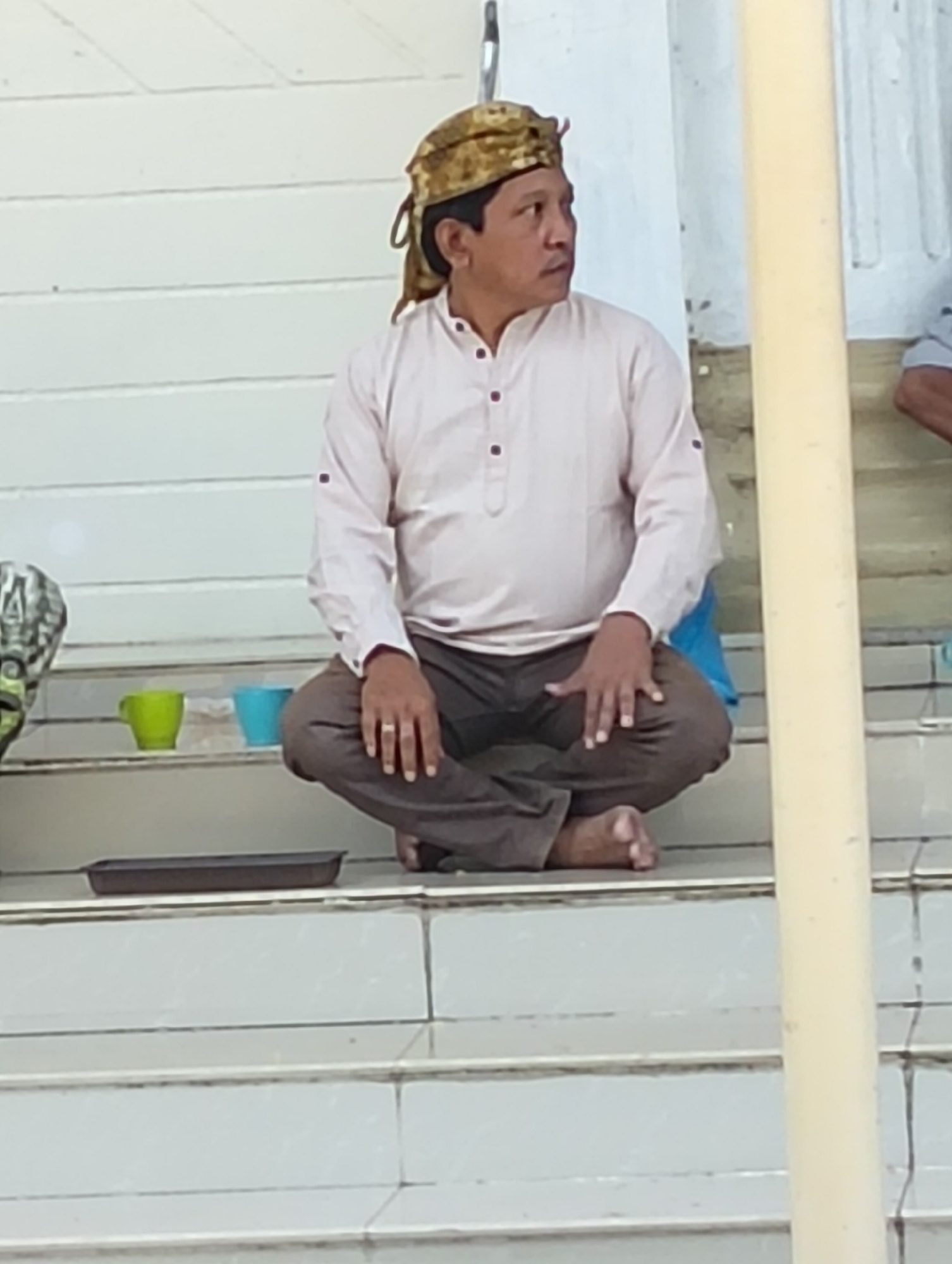 An Indonesian man wearing a patterned head covering, a light-colored collared shirt, and dark trousers sits cross-legged on a light-colored, stepped surface. He is looking to his right. In front of him, on the step, are two small cups, one green and one blue, and a dark rectangular tray. To his right is a thick, pale yellow cylindrical pillar. The background consists of white horizontal planks.