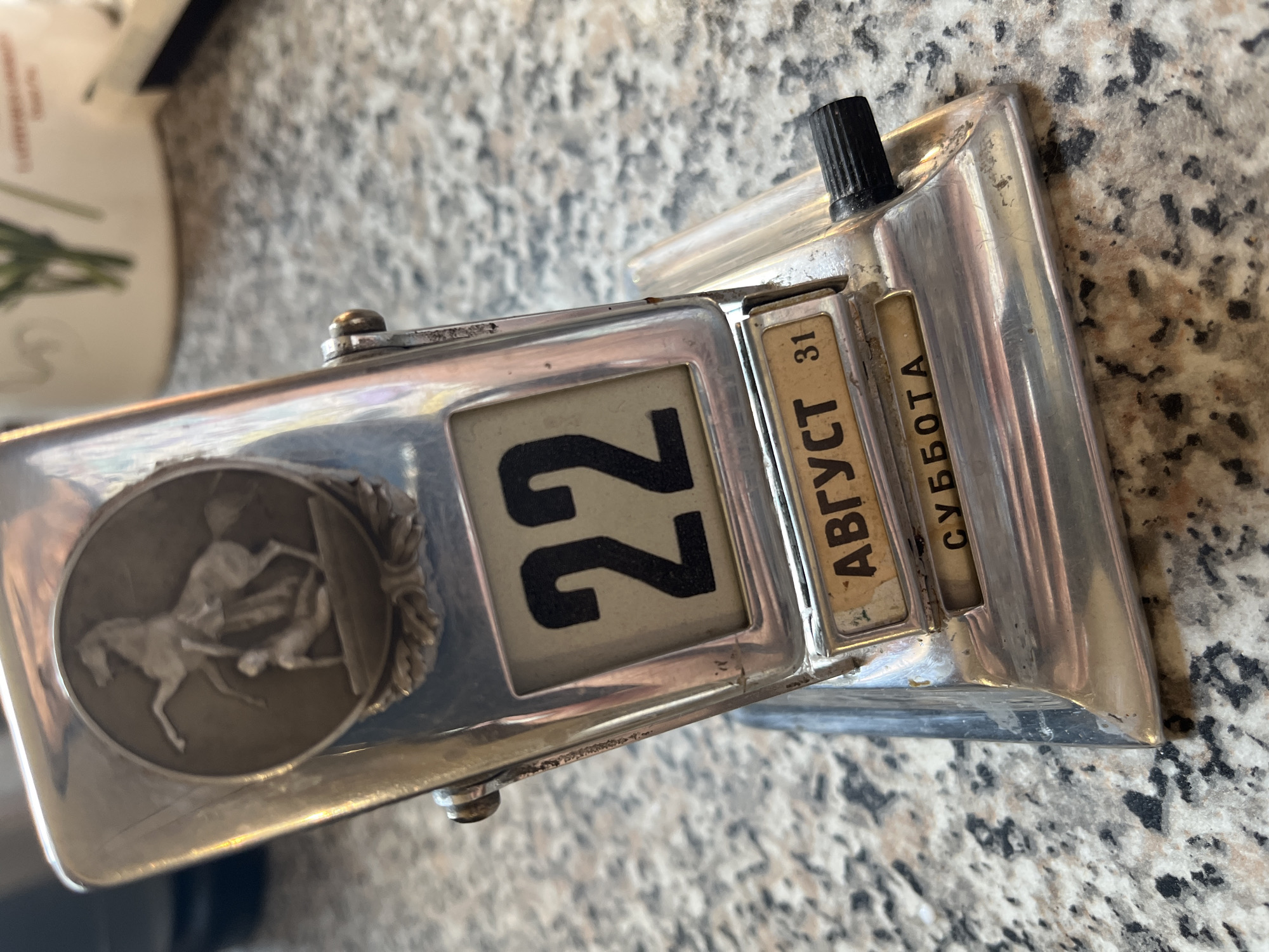 A close-up, eye-level, indoor shot shows a vintage perpetual calendar. The calendar is made of chrome-plated metal and sits on a speckled granite countertop. The calendar has a circular relief on its front depicting a man and a horse. Below the relief, a flip-card display shows the date "22" in black digits on a cream background. Below that, a smaller display shows the month "АВГУСТ" (August) and the number "31" in Cyrillic text. Below the month display, another flip-card shows the word "СУББОТА" (Saturday) in Cyrillic. A black knob is visible on the right side of the calendar, used for changing the date. The background is blurred, showing a white object with green stems, possibly a plant or vase. The lighting is natural, casting a slight sheen on the metal.