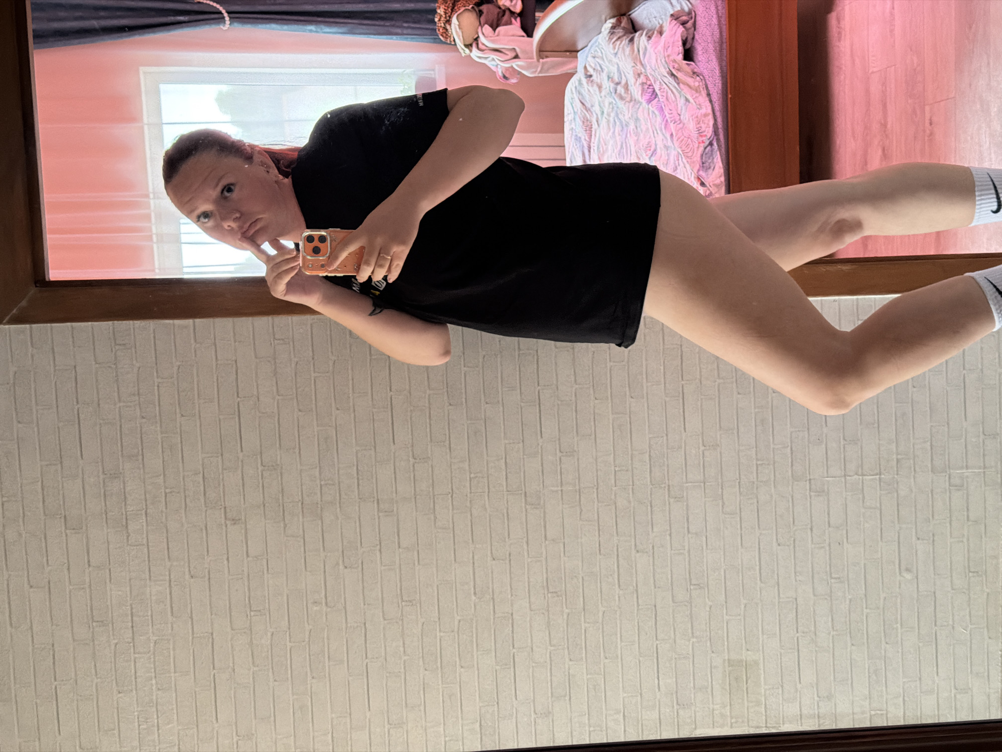 A mirror selfie features a light-skinned individual with reddish hair, posing in a black t-shirt and white socks bearing a black Nike logo. They are holding a phone with a multi-lens camera in their right hand. The reflection shows a room with pink walls, a window with light filtering through, and a bed with a pink and white patterned blanket. The background wall behind the mirror displays a vertical brick pattern. It is not possible to determine if the location is in the city of Chaikovsky, Russia from the information provided.