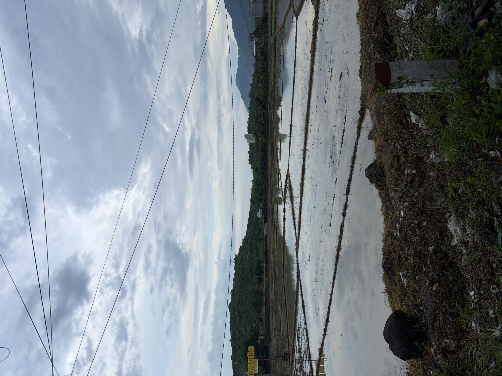 The image depicts a rural landscape in Diên Thọ, Vietnam, characterized by flooded rice paddies reflecting an overcast sky. Power lines stretch across the upper portion of the frame, partially obscuring the sky and the distant, tree-covered hills. In the foreground, the edge of the paddies is lined with dry grass, soil, rocks, and scattered debris including plastic bags. A concrete marker with a red top is situated in the lower right corner, partially overgrown with a vine. A large, dark rock is visible on the left side of the foreground. The overall scene suggests a transitional period, possibly after rainfall, with evidence of human activity in the form of infrastructure and discarded waste, juxtaposed with the natural elements of the agricultural landscape and the surrounding hills.