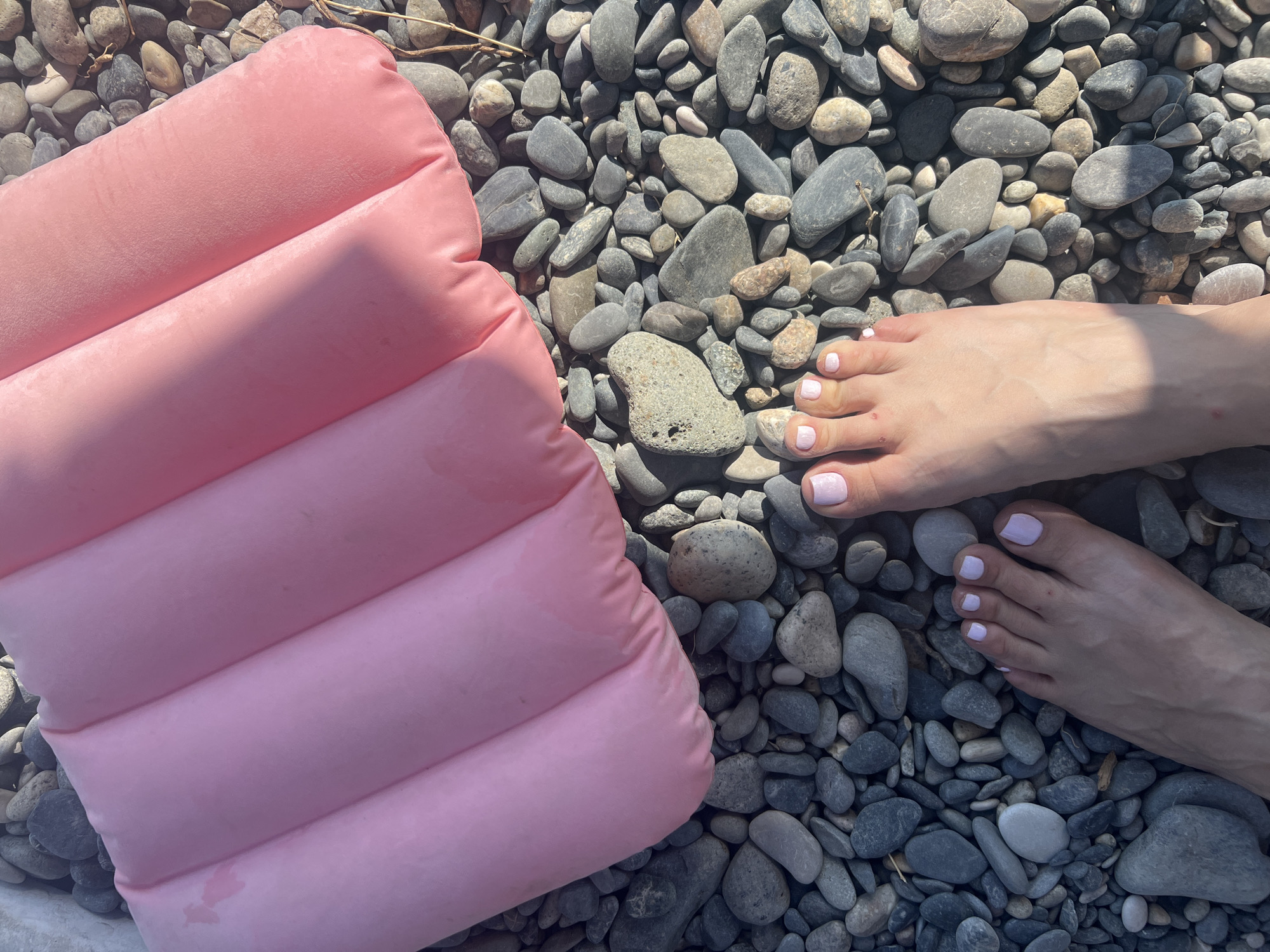 A close-up, overhead shot shows two bare feet, with pale skin and light pink nail polish, positioned on a bed of smooth, grey and brown pebbles. A pink inflatable mat, with a quilted pattern, rests on the pebbles behind the feet. The scene appears to be outdoors, likely on a beach or riverbank, given the pebbles and natural light. The location is specified as Sokhumi, Abkhazia. The overall impression is one of relaxation and leisure.