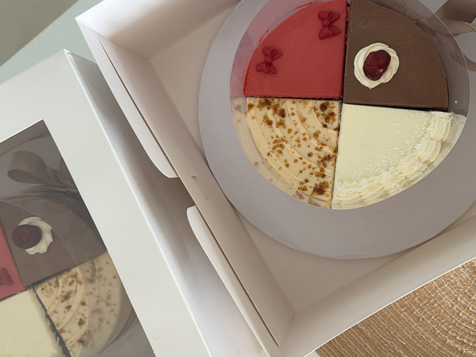 The image displays two white cardboard cake boxes, positioned on a light brown, woven surface. The box in the foreground is open, revealing a circular cake on a white base within a clear plastic liner. This cake is segmented into four distinct quarter-slices. Clockwise from the top-left, the slices are: a light brown or caramel-colored segment with curved ridged patterns and scattered brown speckles; a vibrant red segment adorned with two small red bow-shaped decorations; a dark chocolate-colored segment topped with a dollop of white cream and a single red berry; and a creamy white segment with a ribbed texture along its curved edge and fine white sprinkles. The second box in the background is partially visible, appearing closed with a transparent window on its lid, revealing another similar multi-flavored cake inside, including a dark chocolate quarter with a cream dollop and red berry, and a light-colored quarter. A light brown ribbon is visible within the background box. The scene is uniformly lit, consistent with an indoor environment. The image does not provide information to identify its specific location within Tel-Aviv, Israel. No individuals are present.