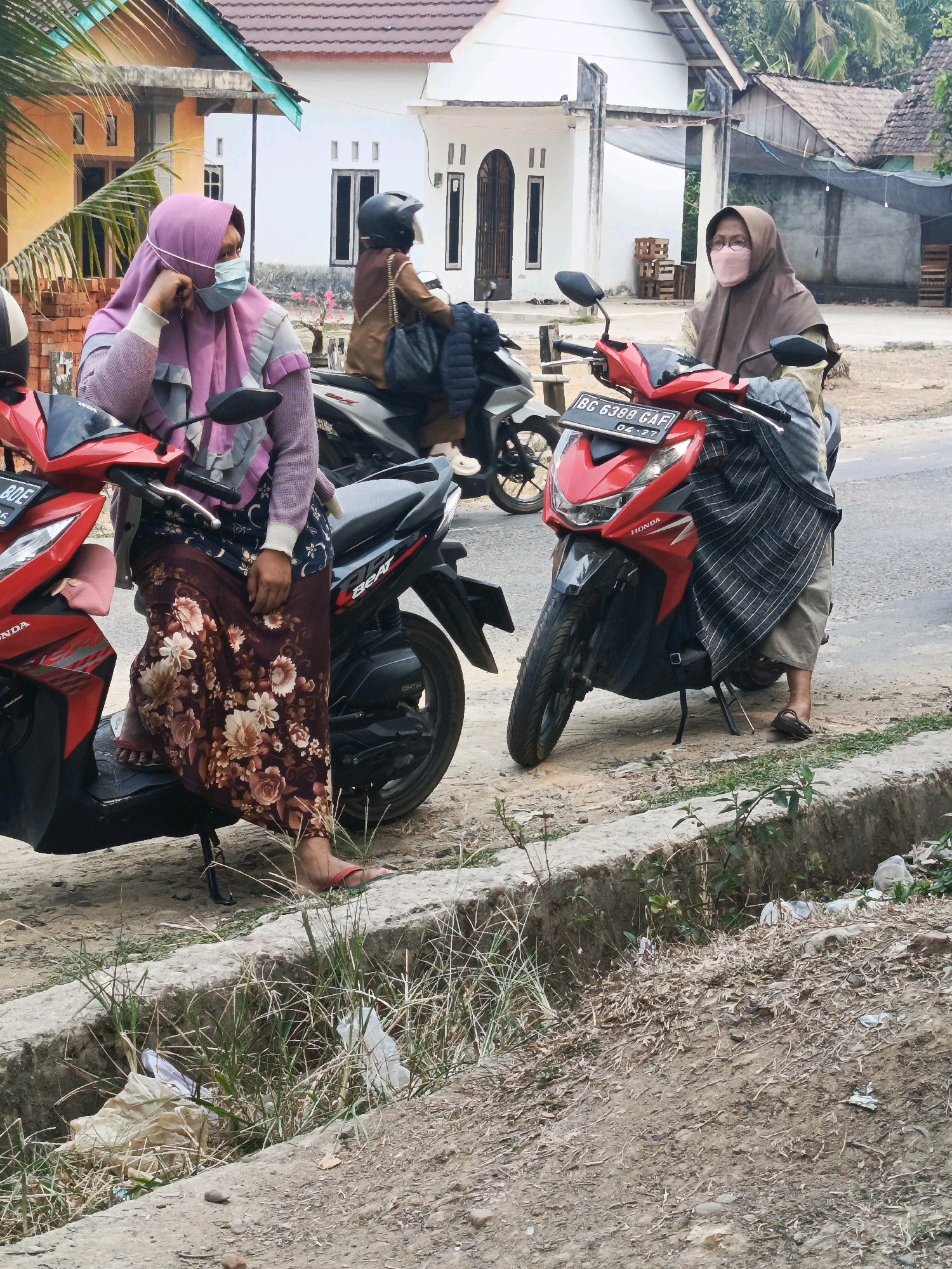 Two women are standing next to motorcycles on a dirt road in front of white houses with brown roofs, likely in Wonosari, Indonesia. The woman on the left is wearing a light purple hijab, a light gray and pink jacket, and a long floral skirt. She has a light blue face mask on and is leaning on her left hand against her head, with her right hand on the motorcycle. The motorcycle is red and black. The woman on the right is wearing a brown hijab and a light-colored shirt with a striped shawl draped over her. She also has a pink face mask on and is standing next to a red motorcycle with the license plate "BG 6388 CAF". The ground is dirt and gravel, with a concrete drainage ditch filled with dry grass and some litter on the right side of the frame. In the background, there are two white houses, one with a teal roof edge, and palm trees. The overall lighting suggests it is daytime.