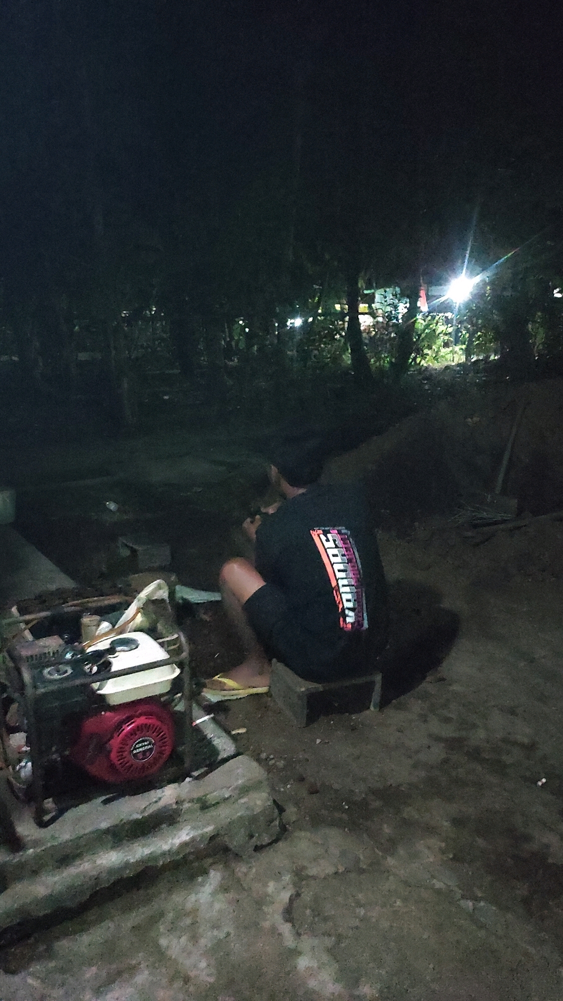 The image depicts an outdoor scene at night in Tanjungan, Indonesia. In the foreground, a red and white portable generator is visible, resting on a cracked concrete surface. To the right of the generator, a person is crouched down, facing away from the camera. They are wearing a black t-shirt with a colorful, abstract graphic on the back and dark shorts. Yellow flip-flops are on their feet. The background consists of dense foliage and trees, illuminated by a bright light source, possibly a lamp, causing a glare. The ground appears to be dirt and loose stones. A shovel leans against a mound of dirt in the far right of the image. The overall ambiance is dim, suggesting it's nighttime in a semi-rural or outdoor area.