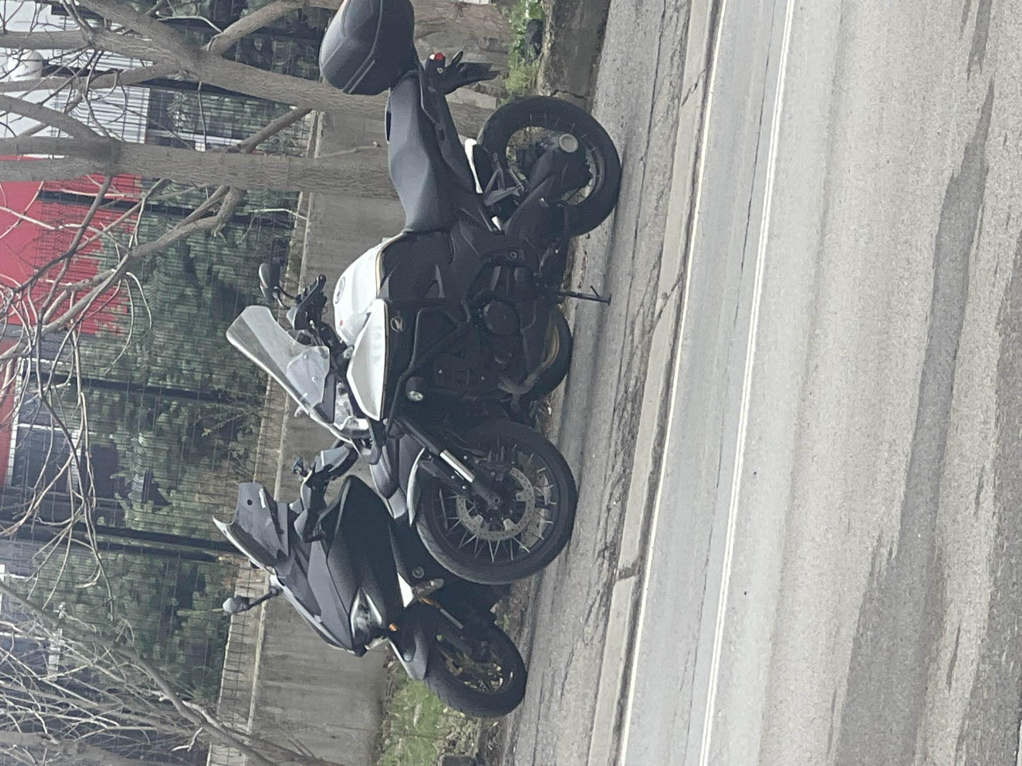 Two motorcycles are parked on the shoulder of an asphalt road. One is predominantly black, the other is black and white. The setting appears to be a city street in Sofia, Bulgaria, based on architectural styles, roadside vegetation, and traffic patterns. The lighting indicates it is daytime. There are trees and a fence behind the motorcycles. The scene is static, with no visible people or immediate activity.