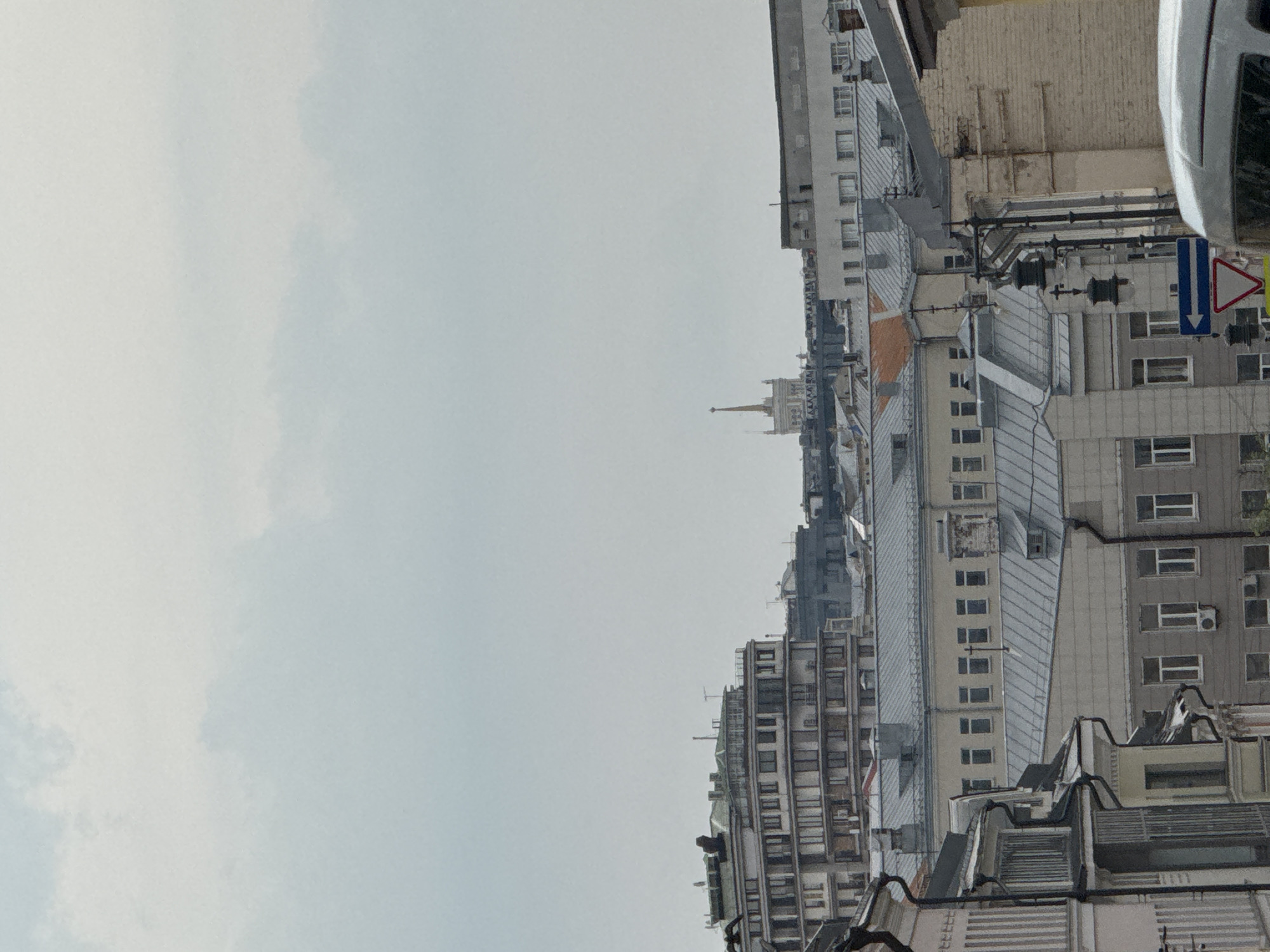This image shows a street view in Moscow, Russia. It features a row of tall buildings with varying architectural styles and heights. The buildings are predominantly white, grey, and beige, with a mix of windows and balconies. The sky is overcast and grey. A spire is visible in the distance. Street signs and electrical wires can be seen, contributing to the urban environment. The photograph is oriented vertically, with the sky occupying the left side of the frame.
