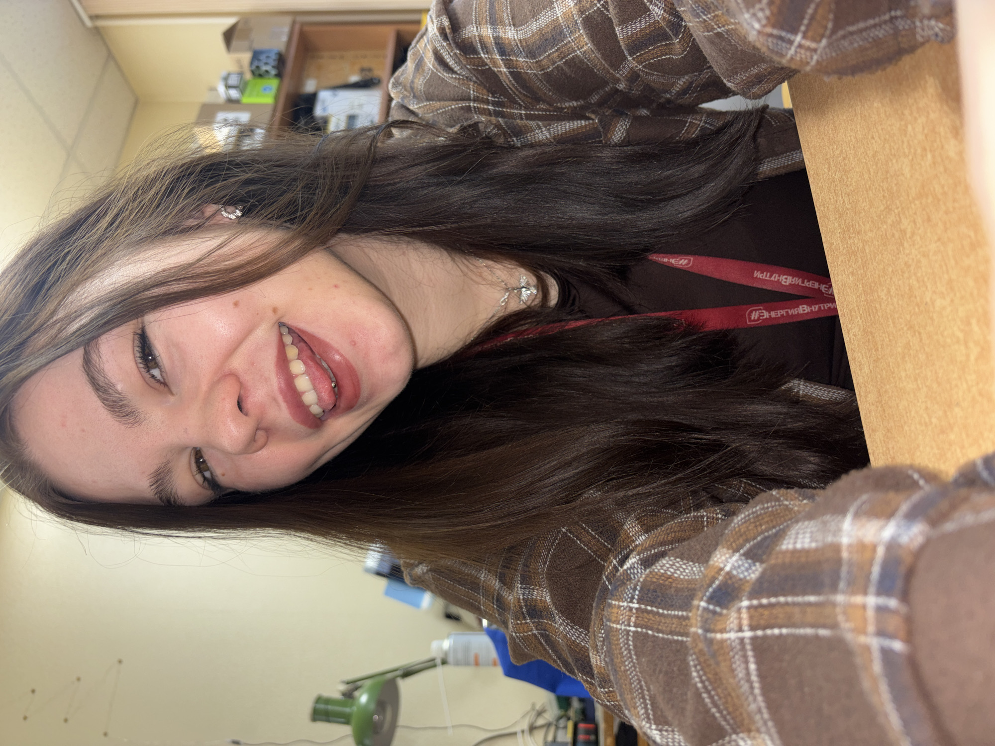 A young woman is taking a selfie, sticking out her tongue, with a table in the foreground. The woman has long brown hair, wears a brown plaid shirt and a dark brown top. She has a tongue piercing and a silver necklace. She is wearing an employee identification tag with the Russian words "#ЭнергияВнутри" (translated as "#EnergyInside") and "ЭНЕРГИЯ ВНУТРИ" (ENERGY INSIDE) printed on the lanyard, indicating she might be an employee. Behind her is a cluttered office area with shelves and boxes. The presence of Russian text suggests the image was taken in Russia. Based on the information on the employee ID tag, it could be assumed she works in the energy field. Without further information, the specific location in the city of Perm is not possible to identify.