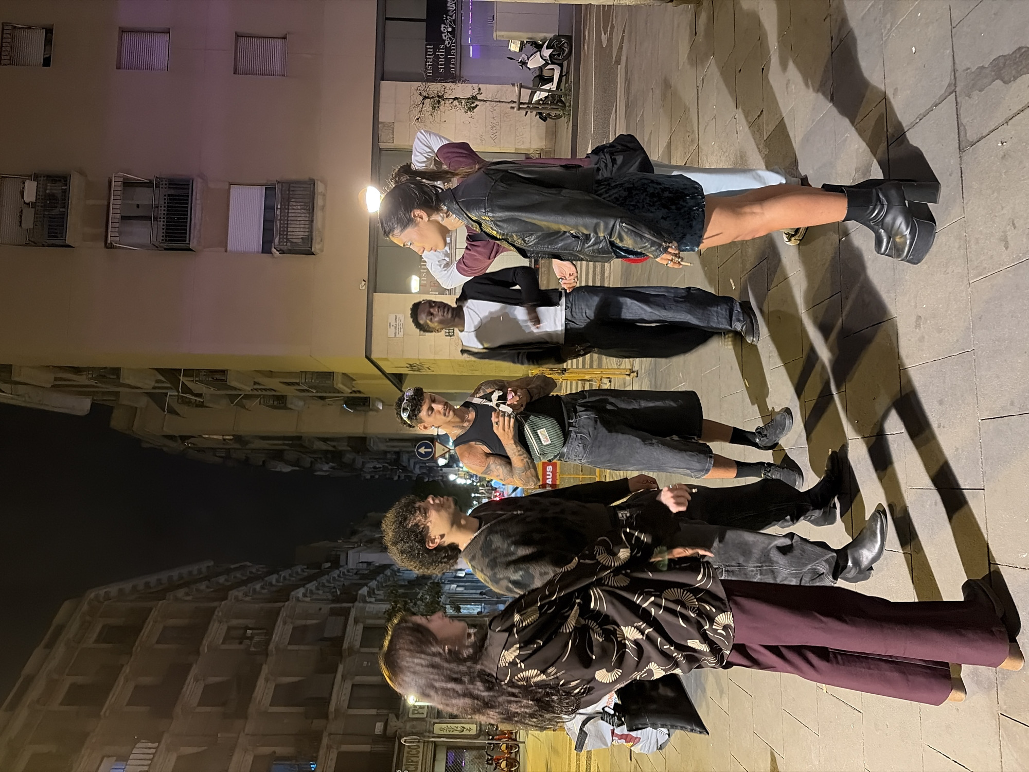 A group of people stand on a sidewalk in Barcelona, Spain. From foreground to background: a woman with long hair wears a patterned top and maroon pants, facing another person with curly hair, wearing a patterned jacket and dark pants. Behind them, a man with tattoos wears a tank top and jean shorts, with a fanny pack. Further back, a man in a white shirt and dark pants stands, followed by a woman wearing a black leather jacket and high-heeled boots. Buildings line the street, and a motorcycle is parked in the background.