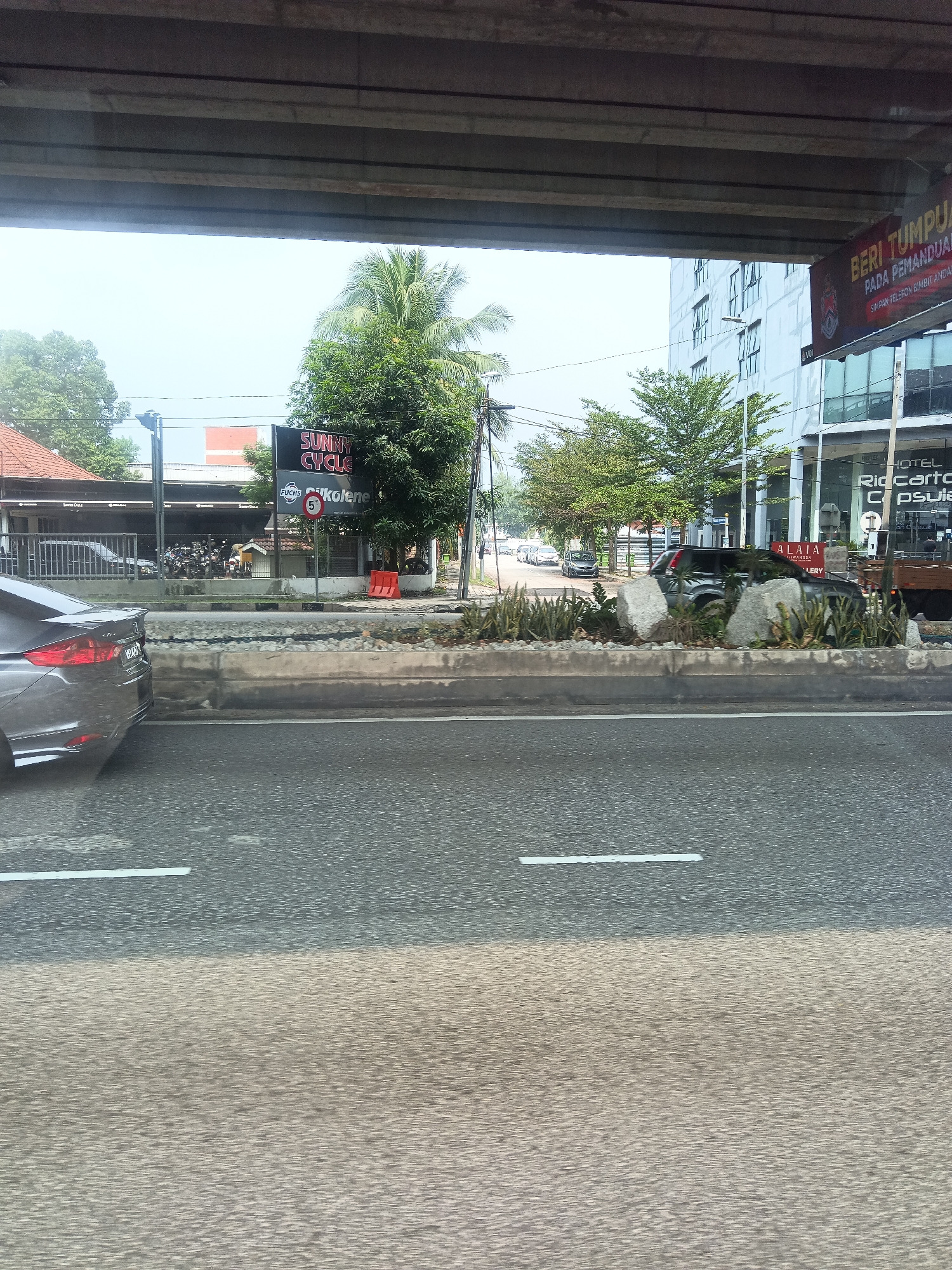 The image shows a street scene in Kuala Lumpur, Malaysia, viewed from a moving vehicle under an overpass. A gray sedan is partially visible on the left side of the frame, its rear end facing the viewer. The road has visible white lane markings. In the background, a cluster of shops and buildings line the street, with a sign for "SUNNY CYCLE" and "FUCHS" prominently displayed. A traffic sign with a "5" inside a red circle indicates a speed limit of 50 km/h. Trees, including a palm tree, are scattered throughout the scene. A modern building with glass windows is visible in the distance. There are no people clearly identifiable in the image. The overall impression is of a bustling urban environment.