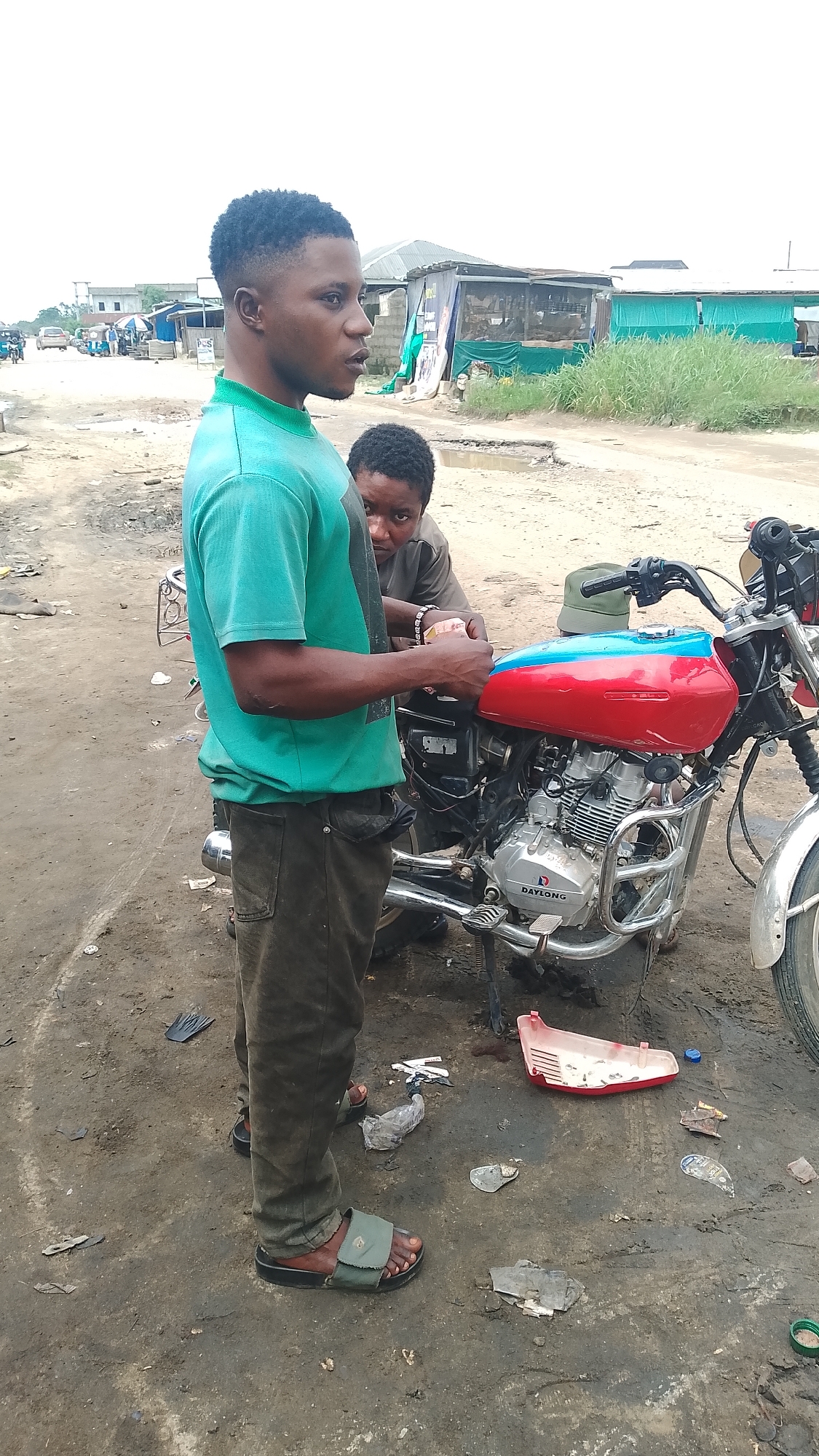 The image depicts a scene on a dirt road, likely in Warri, Nigeria. Two individuals are present, focused on a red and blue motorcycle. The person in the foreground, a young man wearing a teal t-shirt and dark pants, stands looking to the right, his hands near the motorcycle's fuel tank. Another person, whose face is partially visible, is behind the motorcycle, also appearing to be engaged with it. The motorcycle has a chrome engine and a red and blue fuel tank. The environment is characterized by a dusty, uneven ground with scattered debris, including plastic wrappers and a red plastic object. In the background, there are makeshift structures, possibly shops or stalls, and other vehicles, suggesting a commercial or roadside area. The sky is overcast.