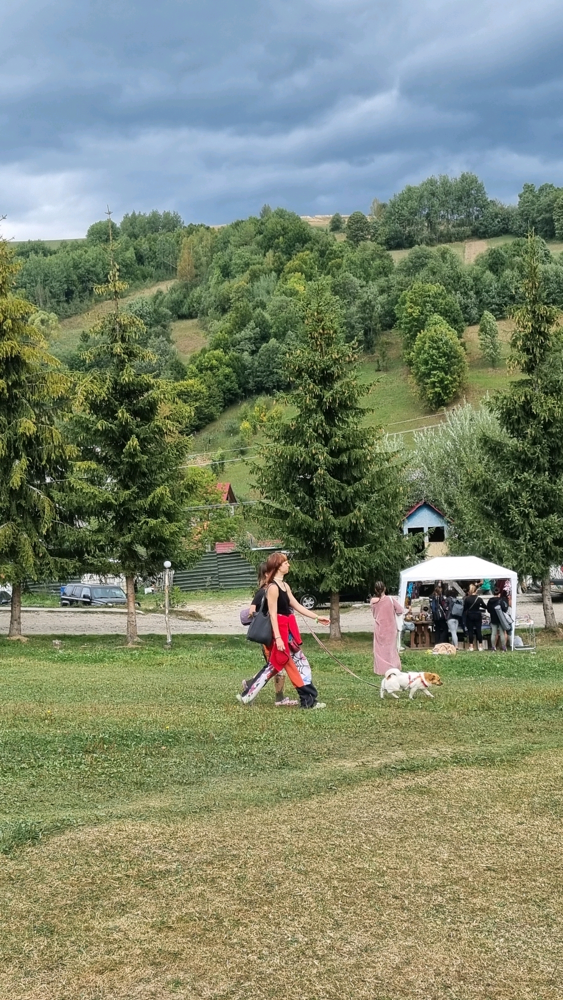 The image depicts an outdoor scene in the village of Izky, Ukraine, under a cloudy sky. In the foreground, a grassy field is visible, with patches of dry grass. Two women and a small dog are walking across the field. One woman, wearing a black tank top and a red jacket, is walking a white and brown dog on a leash. She has a dark bag slung over her shoulder and is wearing colorful pants. Behind her, another woman in a long pink dress is walking. In the middle ground, several tall evergreen trees are planted in a line. Beyond the trees, a white tent is set up, appearing to be a market stall with several people browsing. A small blue and red building is situated behind the tent. In the background, a rolling hillside covered in lush green trees slopes upwards. The overall environment suggests a rural or semi-rural setting, possibly a village fair or market.