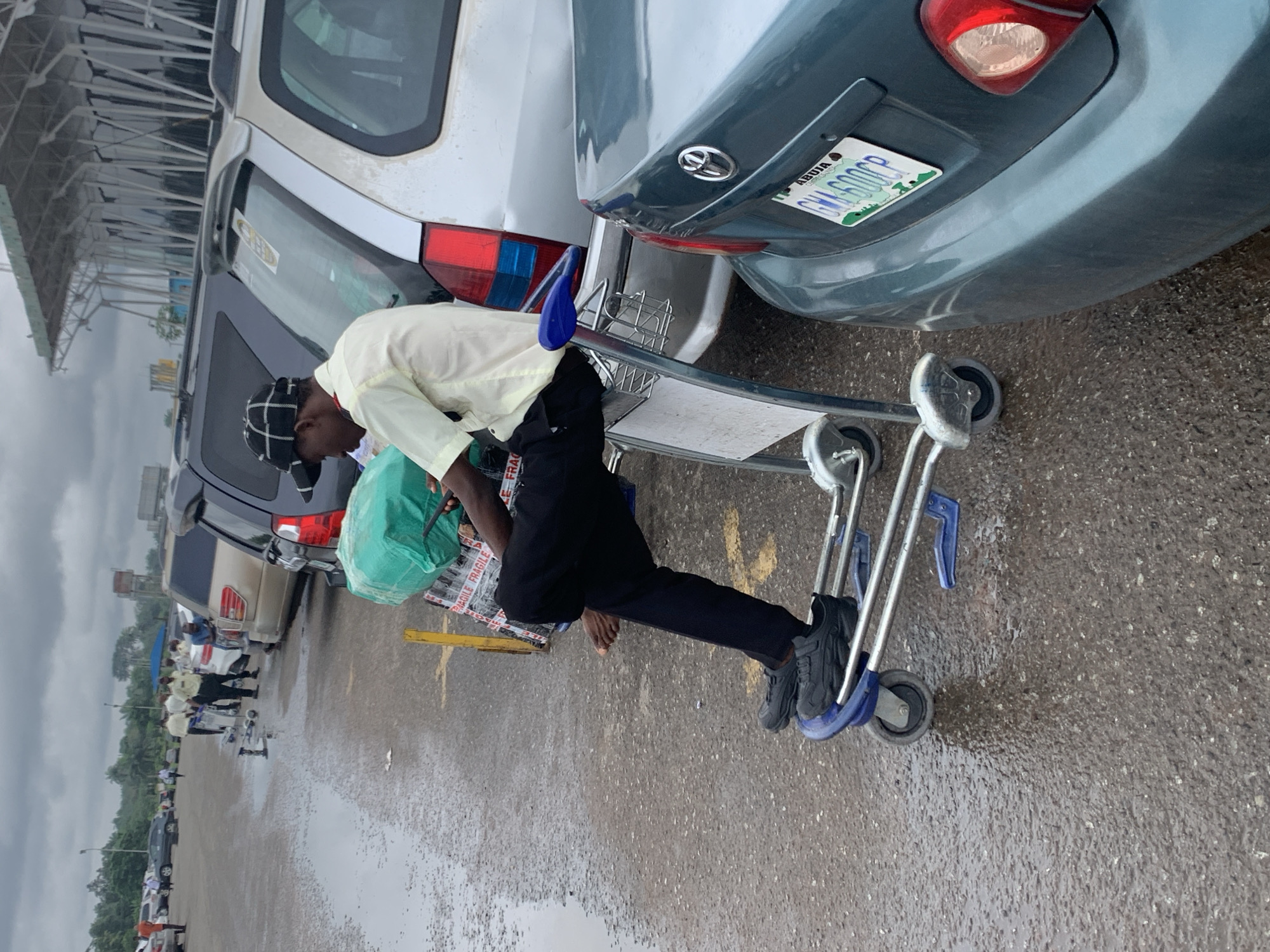 A man sits on a luggage cart with his feet on the bottom rack. He is wearing black pants, a pale yellow shirt, and a black and white checkered cap. He holds a green wrapped package and a phone in his hands. The luggage cart is silver and has four wheels. The man is positioned in a parking lot filled with cars. The ground is wet and reflects the overcast sky. In the background, several other people are visible near other luggage carts and cars. A building with a distinctive white, angled roof structure is visible in the distance. The license plate on the rear of the teal car in the foreground reads "ABUJA GMA 600CP". The scene suggests an airport or transportation hub setting.