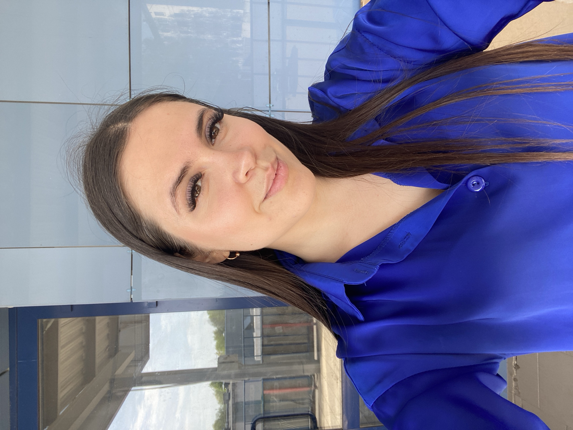 A woman with long brown hair and extended eyelashes is wearing a blue button-up shirt. She appears to be indoors in a building with windows, possibly a train station or bus terminal, based on the visible infrastructure outside the glass. The location may be the city of Chaikovsky, Russia.