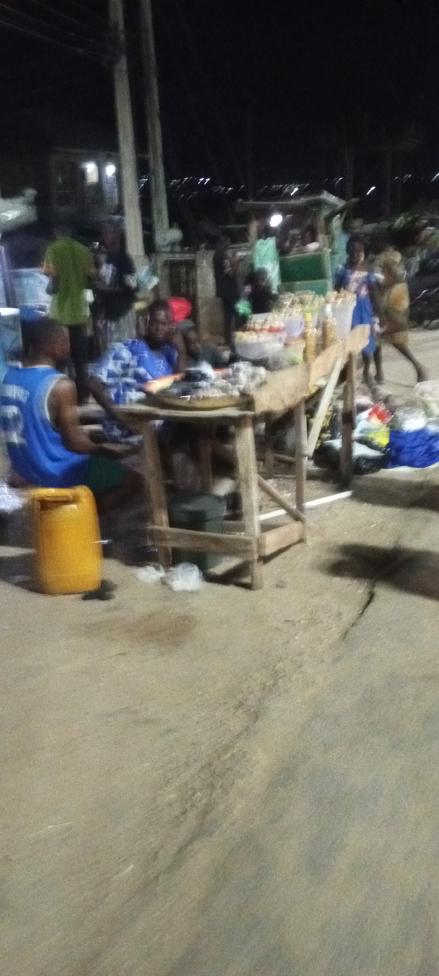 The image shows a nighttime scene in what appears to be a market or street vendor area in Adankolo, Nigeria. A person wearing a blue sleeveless jersey with "32" on the back is seated to the left, facing away from the camera. Next to them is a large yellow plastic container. In the center, another person in a blue and white patterned shirt is seated at a wooden table. On the table, there are various food items displayed, including what appears to be fish on a circular platter. In the background, several other people are visible, along with market stalls, some with goods on display. The lighting suggests it is after dark, with artificial lights illuminating the area. The ground is a paved or dirt surface. The overall impression is of a bustling evening market environment.