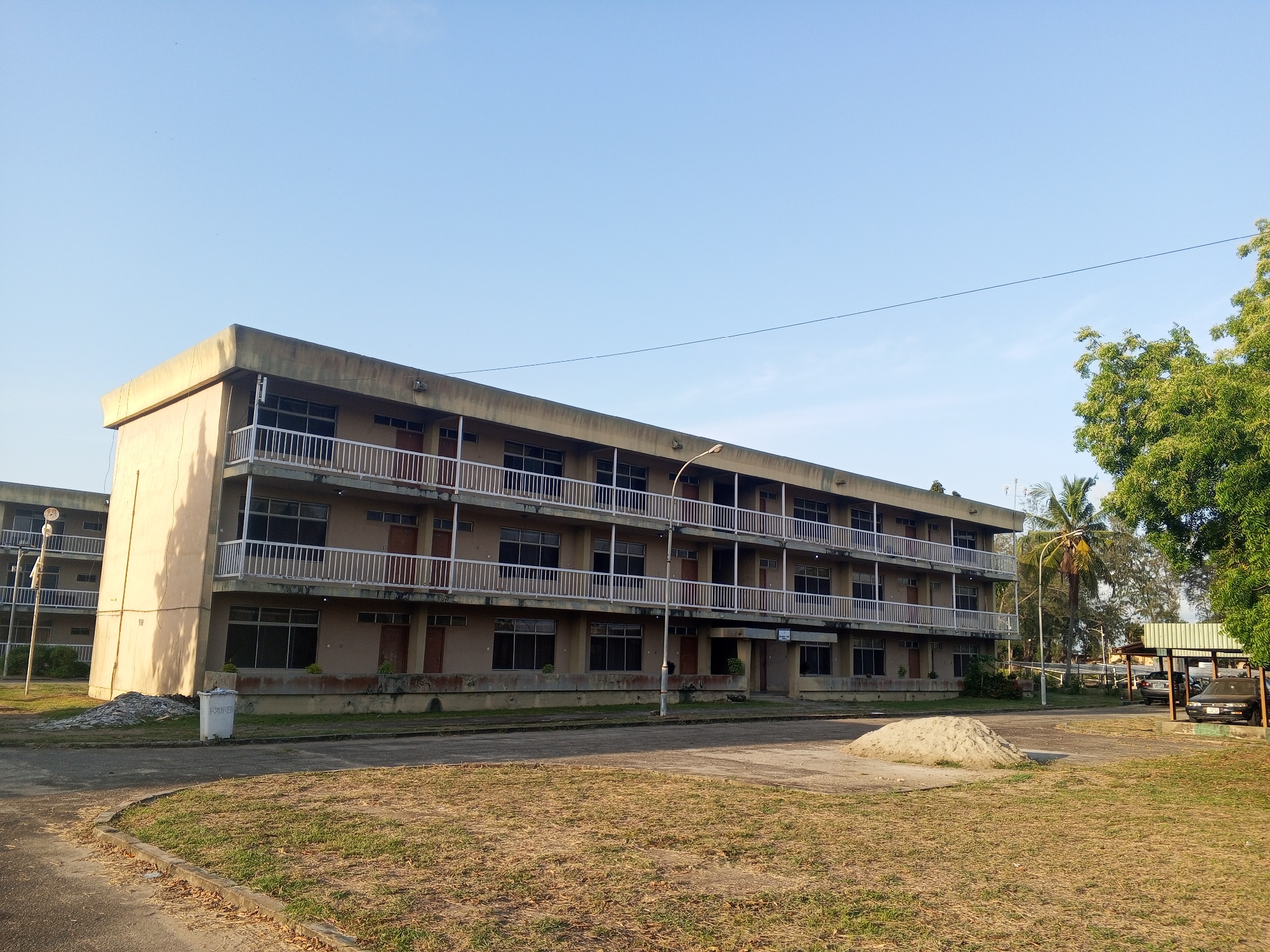 The image depicts a three-story, rectangular building with a flat roof in Tawpo, Nigeria. The building has a beige exterior with white railings on the balconies. There are numerous windows and doors visible on each floor. A street light stands in front of the building. In the foreground, there is a grassy area and a pile of sand. A parking area with a few vehicles is visible to the right of the building, under a metal structure. The sky is clear and blue. No people are visible.