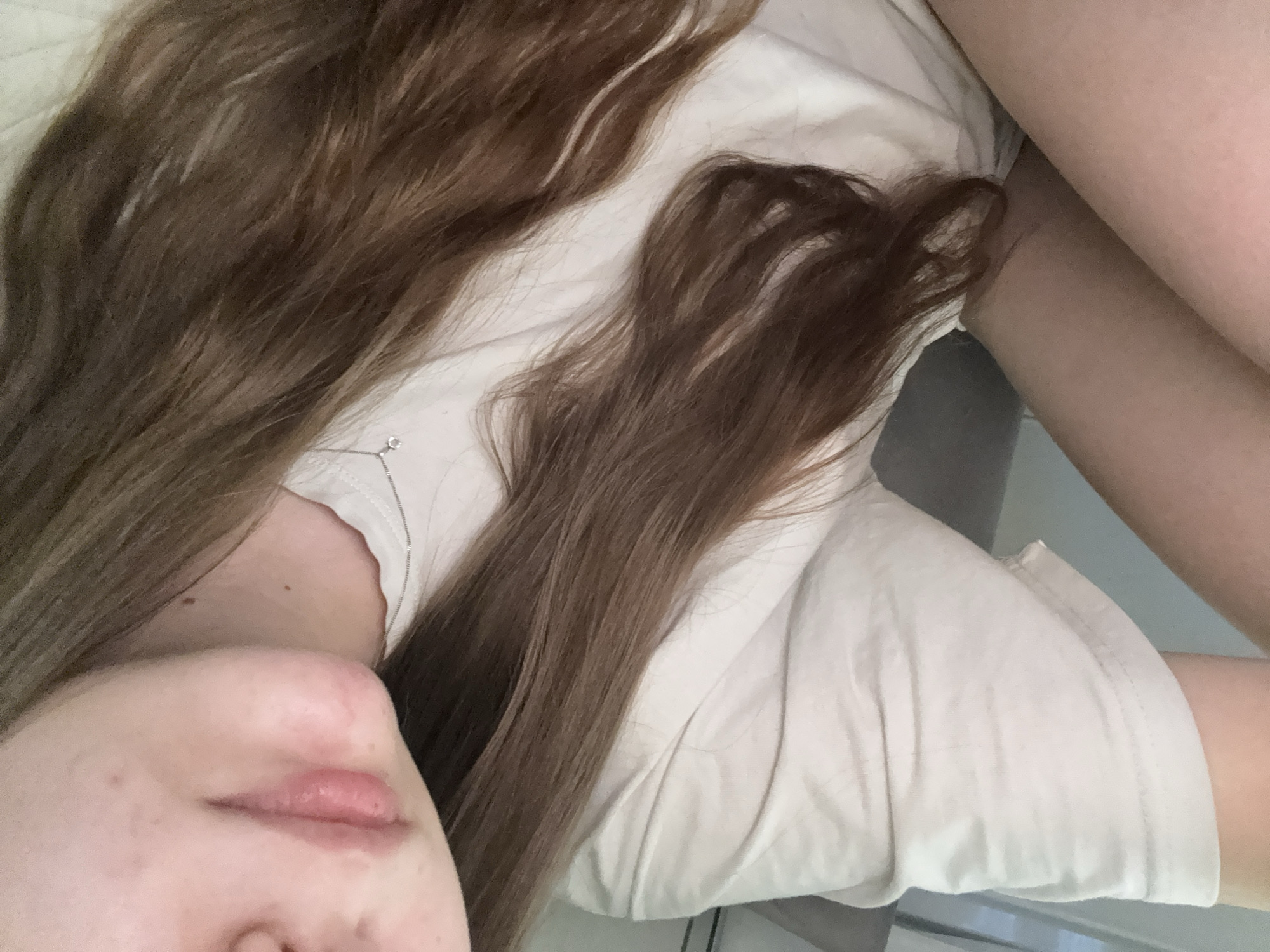 The image presents a close-up, high-angle view of a person, likely female, lying down indoors. The frame captures the lower part of their face, including the chin, mouth, and a portion of the nose and cheek, alongside their shoulder, upper chest, and part of an arm.
The person has long, flowing brown hair with subtle lighter streaks, which is spread across a light-colored surface, possibly a bed or pillow. They are wearing a simple, light-colored (appearing off-white or cream) short-sleeved t-shirt. A delicate silver chain necklace with a small, clear pendant is visible around their neck.
The visible facial skin is fair, with a small mole or freckle present on the lower cheek/chin area. The lips appear naturally colored and slightly full. The posture and composition suggest a moment of rest or relaxation. The lighting is soft and ambient, characteristic of an indoor setting. There are no specific visual cues within the image to indicate the exact time of day or the provided location of Saint Petersburg, Russia. No text is visible.