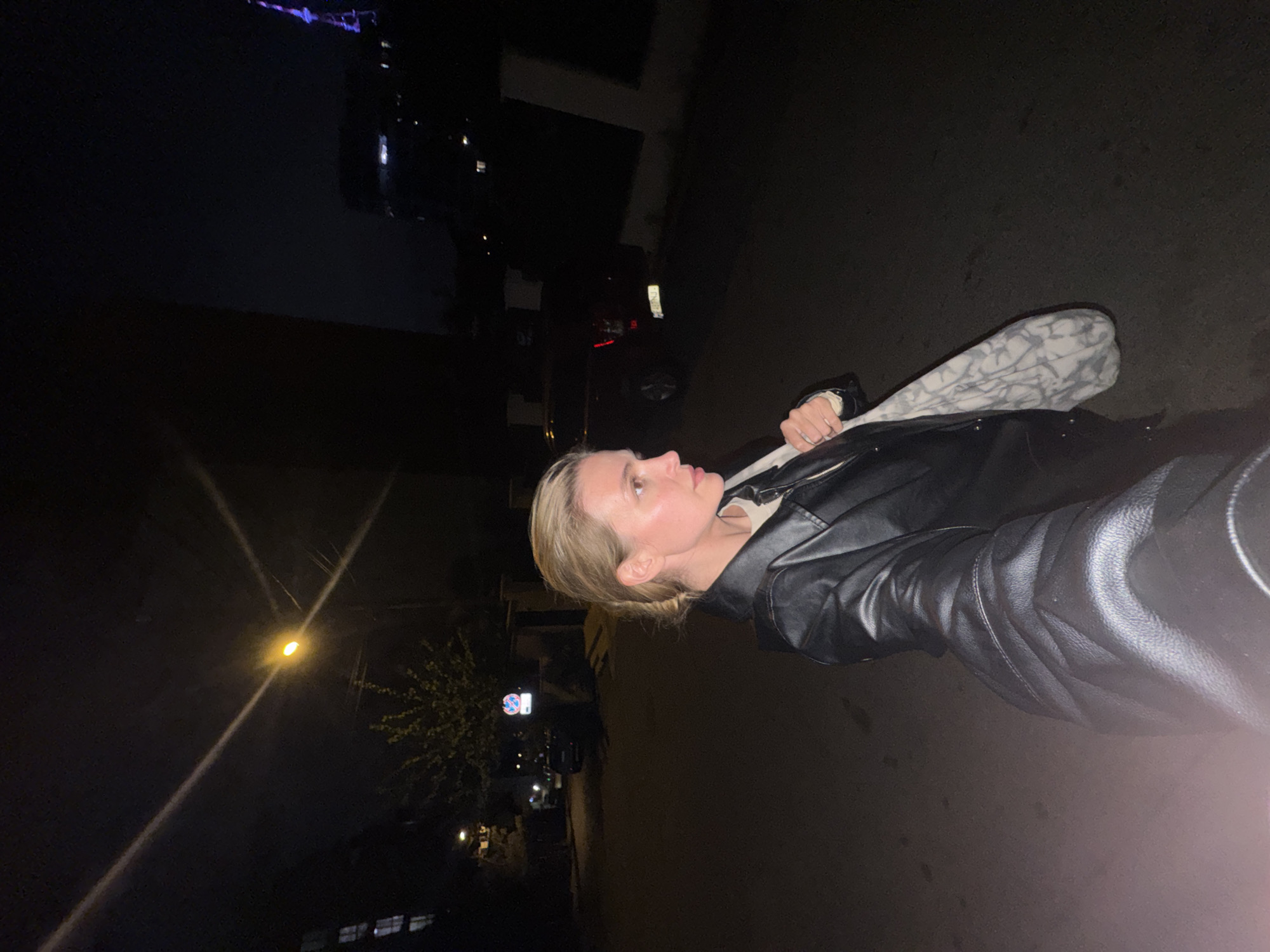 A medium shot photograph depicts a blonde woman, viewed in profile and partially facing away from the camera. She is wearing a black leather jacket and carrying a light-colored patterned bag on her shoulder. The setting appears to be outdoors at night in a city. Street lights illuminate the area around her. In the background, there is a car and buildings with lit windows. Signage in Georgian identifies this location as Tbilisi, Georgia.