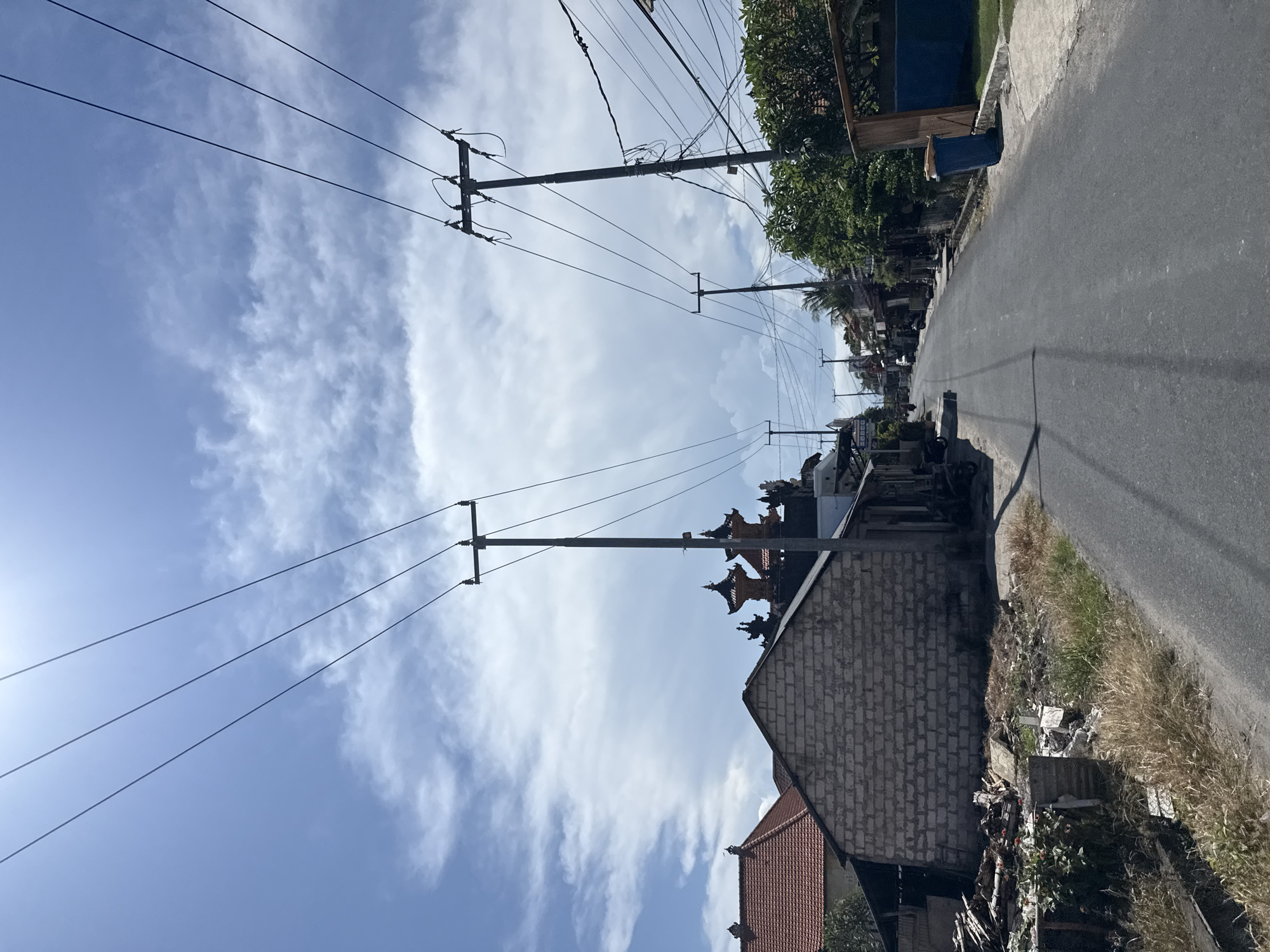 The image shows a street scene in Batununggal, Indonesia. A paved road runs along the right side of the image. Electrical poles line the street on both sides, with power lines stretching across the sky. A building with a brick wall and Balinese architectural details is visible on the right. The sky is blue with scattered white clouds. Vegetation is present along the road's edge. Debris is visible near the building.