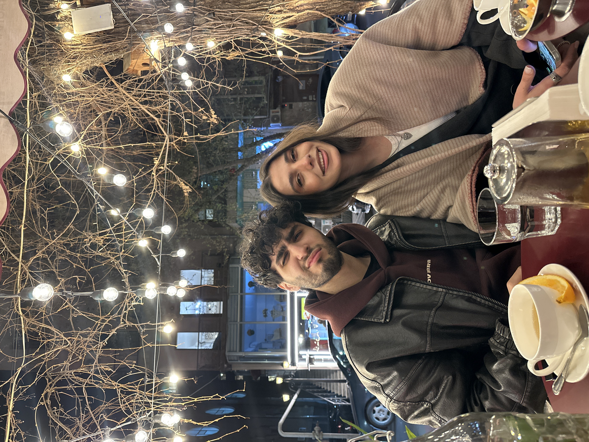 A photo taken at an outdoor cafe in Yerevan, Armenia, shows a woman and a man seated at a table. The woman, with long brown hair, is smiling. She is wearing a light brown shawl and a white top. The man, with dark curly hair and a beard, is making a funny face. He is wearing a brown hoodie under a black leather jacket.
On the table, there is a tea set with a teacup and a glass teapot. The background shows buildings and street lights, indicating an urban setting at night. Above the table, there are decorative bare branches adorned with numerous small string lights, creating a warm, inviting atmosphere.
