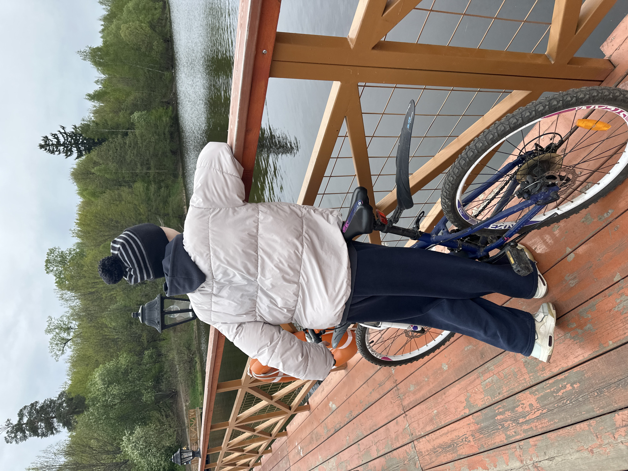 A person stands on a wooden platform, leaning against a wooden railing, with a bicycle beside them. The platform appears to be a pier or walkway extending over a body of water. The person is wearing a light-colored puffer jacket, dark pants, and white sneakers, with a striped hat. The bicycle is blue and white. In the background is a lake or river surrounded by trees. Street lamps are visible along the railing. The location is likely in the city of Grebnevo, Russia.