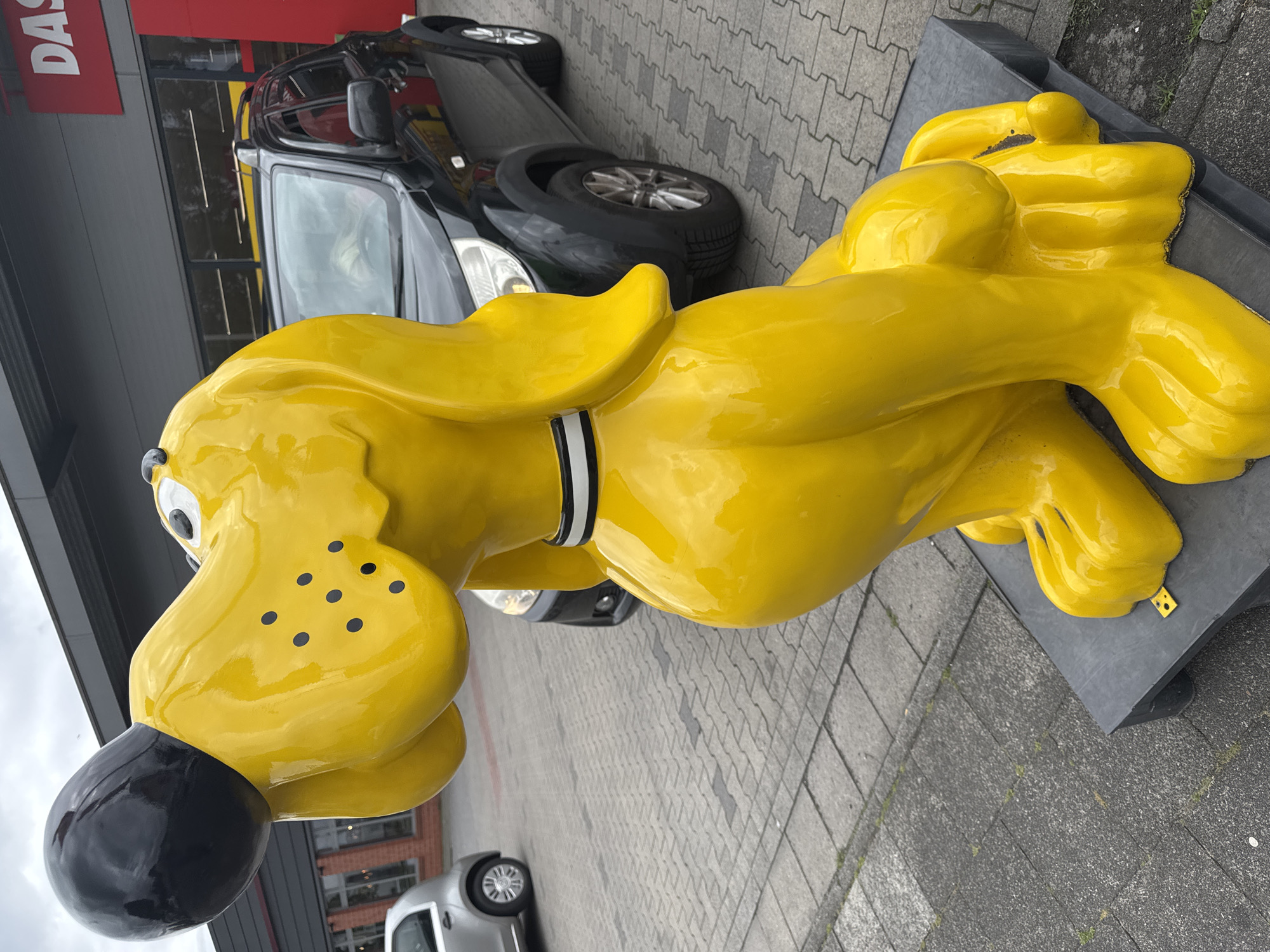 A bright yellow, cartoonish dog statue sits on a gray platform in front of a building with a red sign that reads "DAS". A black SUV is parked behind the statue, and a silver car is visible to the left. The ground is paved with gray rectangular tiles. The sky is overcast. The statue is in Gronau, Germany, at the location of a former "DAS"-branded electronics store.