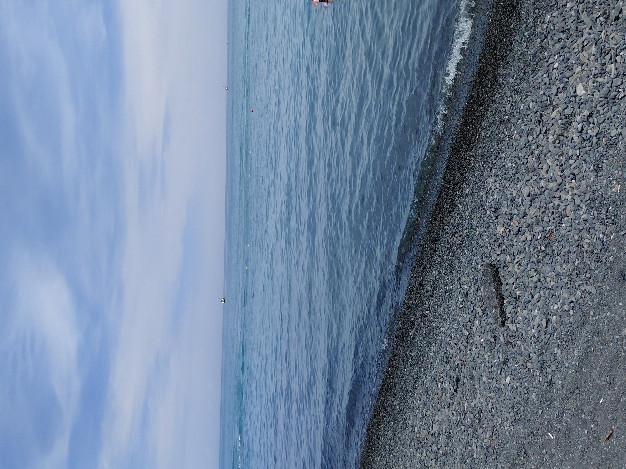 A wide shot captures a pebble beach in Ақши, Kazakhstan, meeting the calm, blue waters of a large body of water. The sky above is a pale blue with scattered, wispy white clouds. The beach is composed of numerous small, gray pebbles, with a few larger, darker stones and a patch of asphalt near the foreground. The water exhibits gentle ripples and a slight wave breaks at the shoreline. In the distance, two small boats are visible on the water, and a person can be seen partially submerged in the water on the right side of the frame. A few orange buoys are also dotted across the surface of the water.