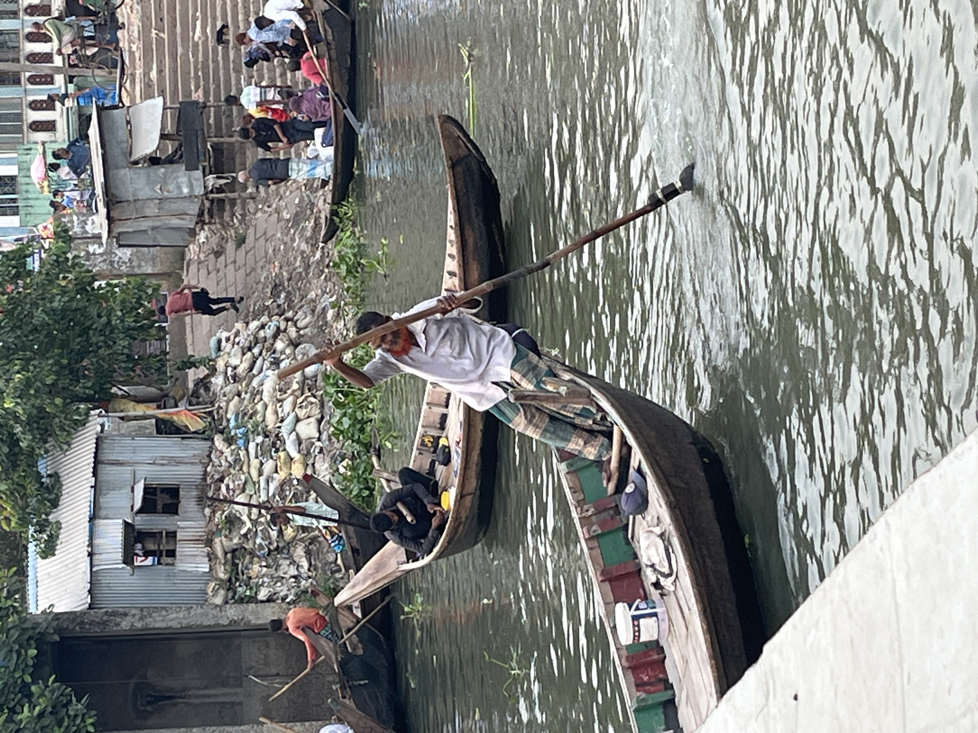 A man in a white shirt and checkered pants stands on a narrow wooden boat in the Buriganga River, propelling the boat with a long pole. Nearby, a similar boat carries a person in dark clothing. Along the riverbank in Dhaka, Bangladesh, are buildings with corrugated metal roofs, piles of garbage, and people moving about. The river reflects the light, creating a rippled pattern on the water's surface.