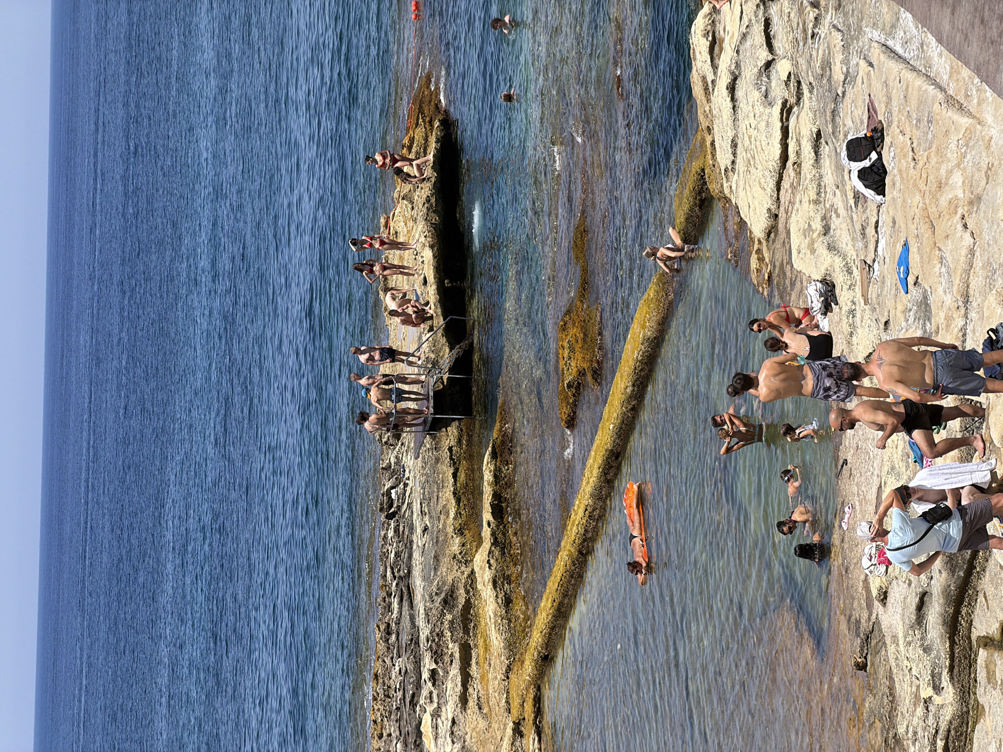 The image depicts a sunny day at the rocky coastline of Sliema, Malta, where numerous people are enjoying the natural rock pools and the clear blue sea. In the foreground, several individuals are situated on the rocky shore, some in swimwear. A man in a blue t-shirt and grey shorts stands facing away from the camera, adjusting his hat. Beside him, another man sits with a towel draped over his shoulder, and a third man in patterned shorts stands facing the water.
In the shallow, clear rock pool, a woman is seen relaxing on an orange inflatable mat. Further into the pool, groups of people are swimming and interacting. To the right of the pool, a raised rock platform juts out into the sea. Several people are standing and sitting on this platform, also enjoying the sun and the water. Some are dressed in bikinis and swim trunks, while others are bare-chested.
The environment is characterized by sun-drenched, uneven rock formations and the tranquil, deep blue of the Mediterranean Sea. The water is clear, revealing the submerged rocks and the seabed in places. The sky above is a clear, bright blue, indicating fair weather. The overall scene conveys a sense of leisurely summer activity at a popular coastal spot.