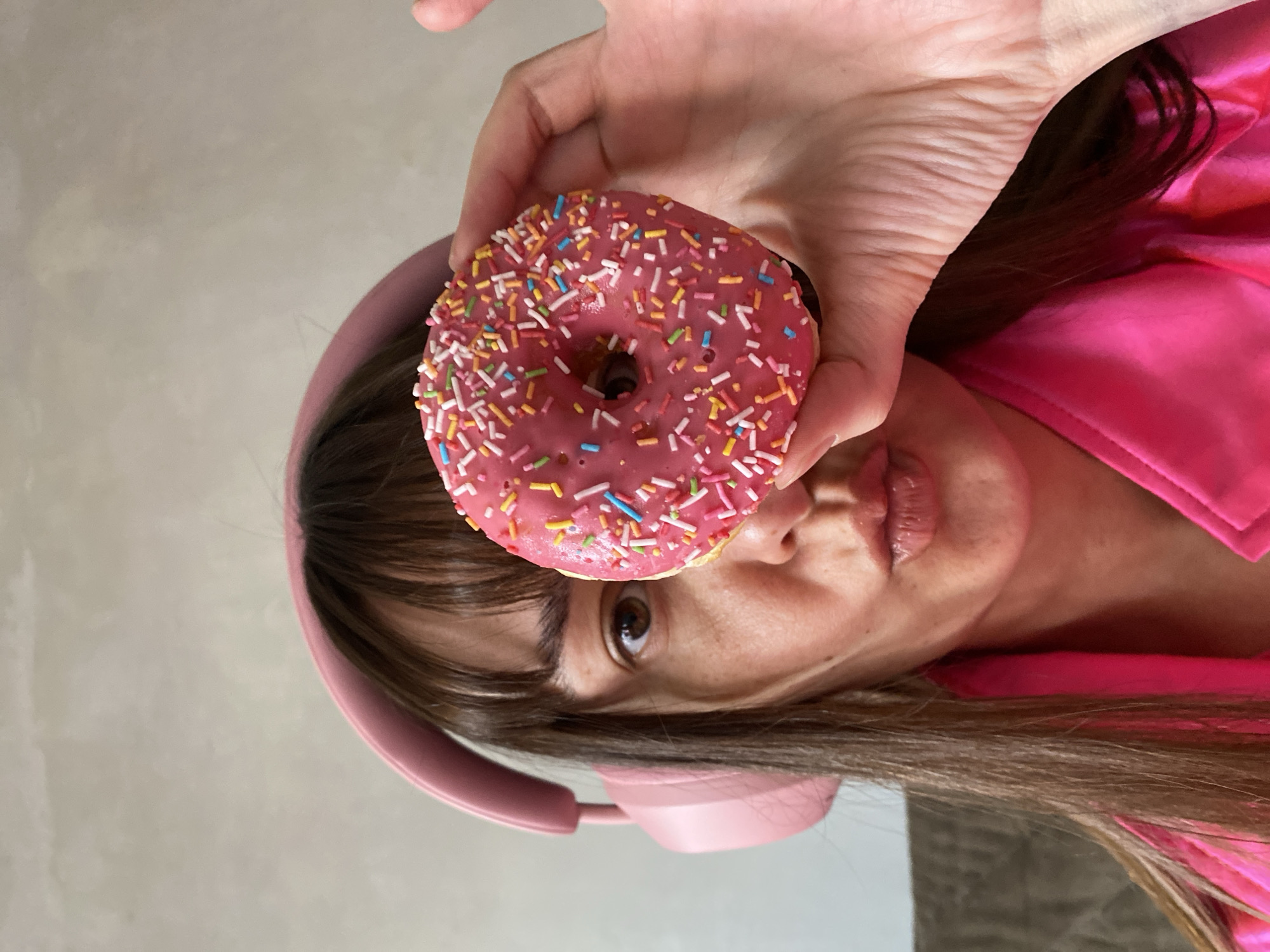 An expressive young person, likely female, is captured in a close-up, rotated portrait, seemingly indoors in Votkinsk, Russia. Her head is tilted, giving the impression that she is looking upward and to the side. She has medium-brown hair with bangs and is wearing pink over-ear headphones around her neck or ears, matching her bright pink satin top.
The central focus of the image is a vibrant pink-frosted donut, generously covered in a variety of colorful sprinkles (pink, yellow, blue, white, orange). The donut is held aloft by a hand, positioned just above and slightly in front of her face, obscuring part of her forehead and hair. Her right eye is clearly visible, dark brown, and looks directly towards the camera, conveying a playful or mischievous expression. Her lips are slightly parted, revealing no teeth. The background is a plain, light, neutral-colored wall, offering no additional context regarding the specific location or time of day. The overall impression is one of lightheartedness and a focus on the colorful, sweet treat. No visible text is present in the image.