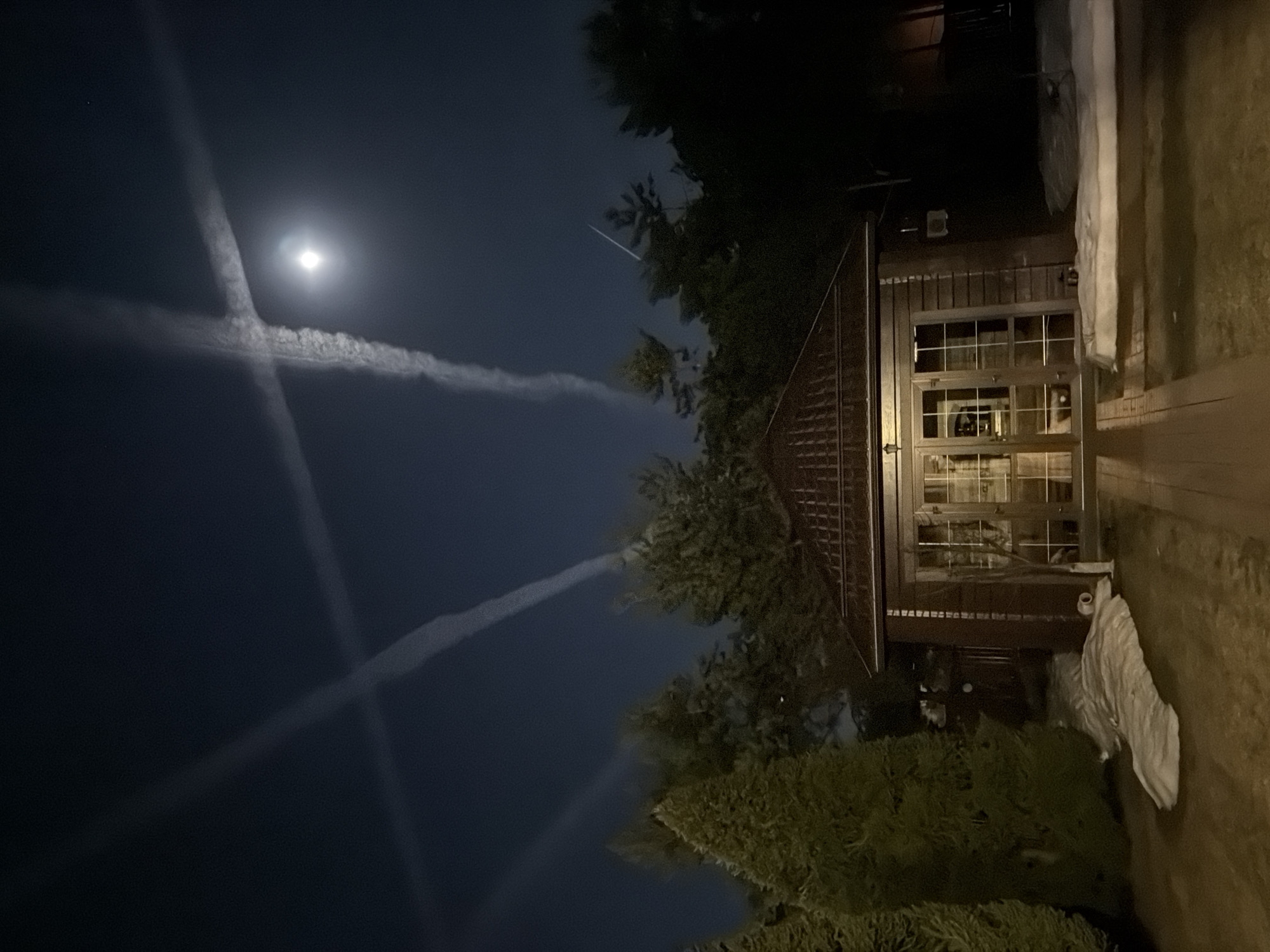 The image shows a nighttime scene. A small wooden building with a multi-paned window and a gabled roof is visible. The building is situated amongst trees. The sky is dark with what appears to be contrails intersecting near a bright moon. A possible meteor streak is visible to the right of the moon. The photo is taken in Mishutkino, Russia. No people are visible.