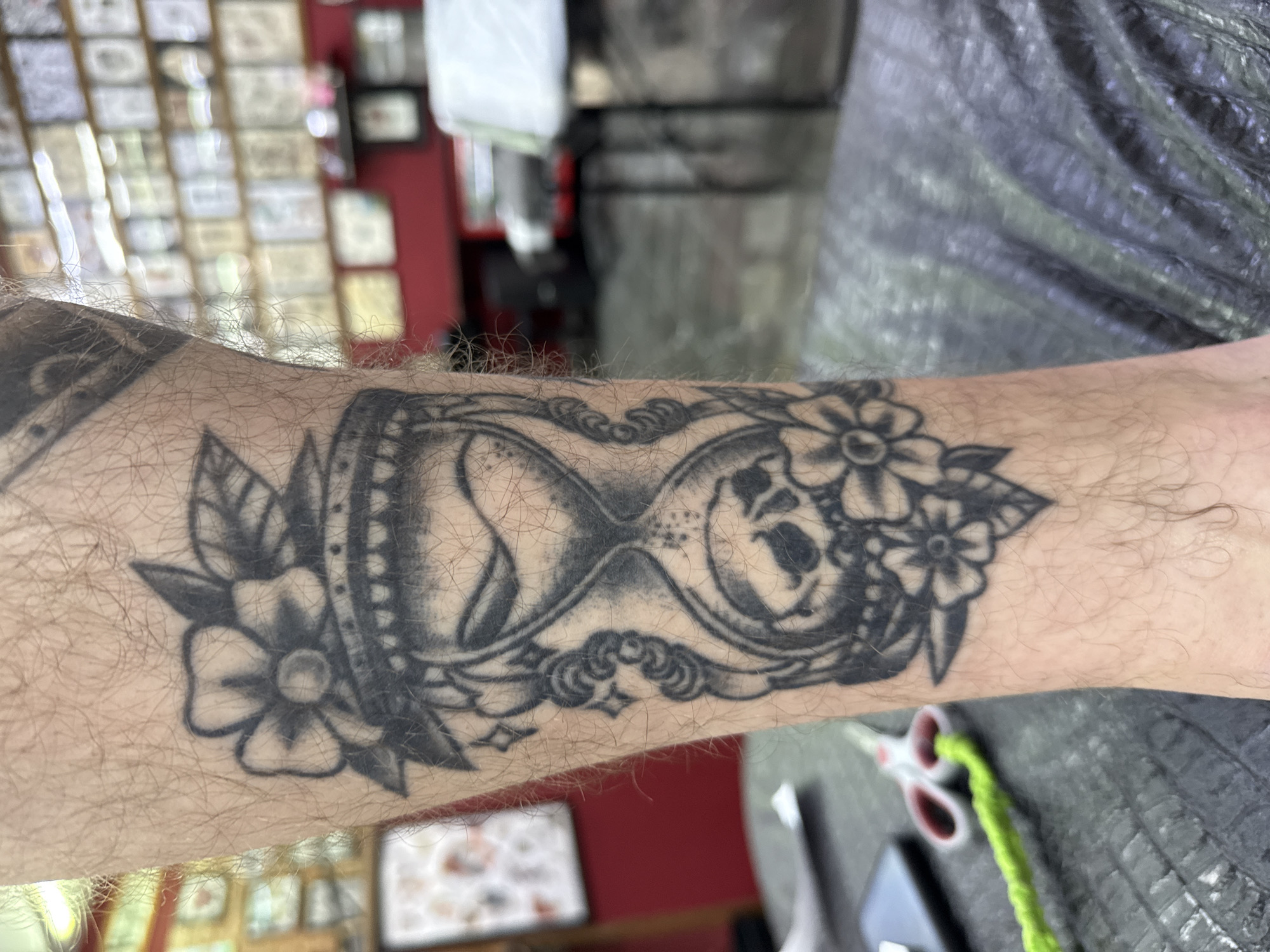 The image shows a close-up of a tattoo on a person's leg. The tattoo is a black and grey hourglass with a skull inside. It is surrounded by flowers and leaves. The background is blurred and appears to be a tattoo parlor, with walls covered in tattoo designs and a table with scissors and a phone. There is no information about the location in Moscow, Russia.