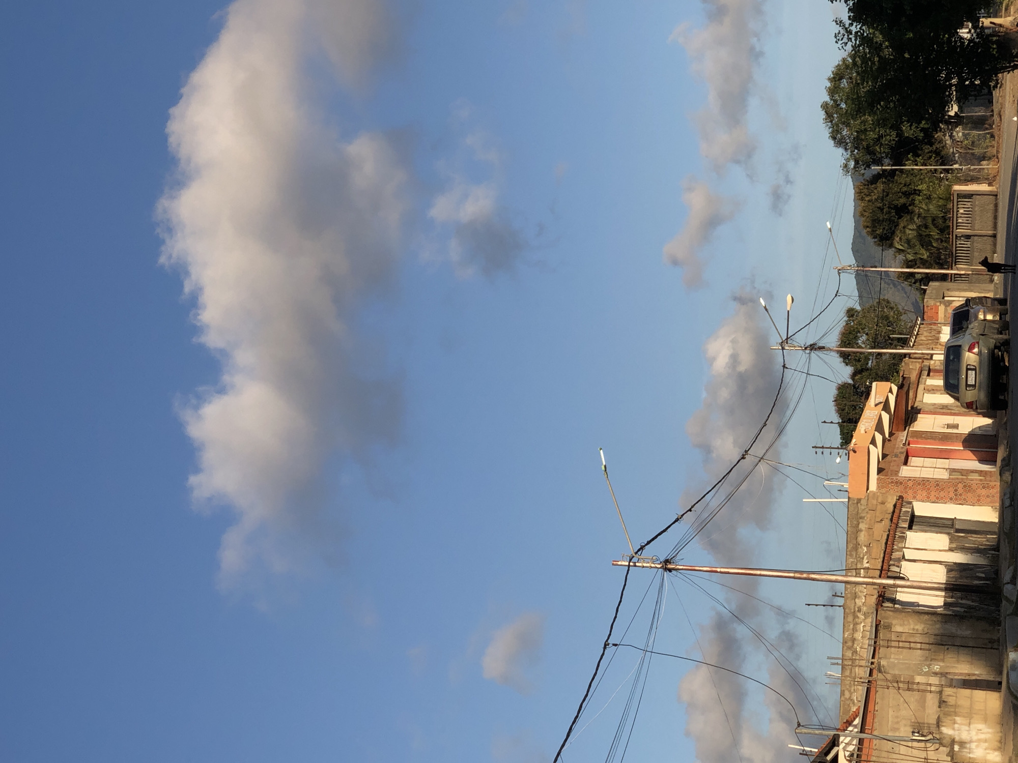 The image depicts a street scene in Altagracia, Venezuela. A large white cloud dominates the left side of the sky. On the right, the sky is blue with smaller clouds. Buildings, power lines, and streetlights are visible on the right side of the image. A dark-colored car is parked along the street, and a dog stands near the edge of the road. In the background, a mountain is partially obscured by trees. The scene appears to be captured during daylight hours.