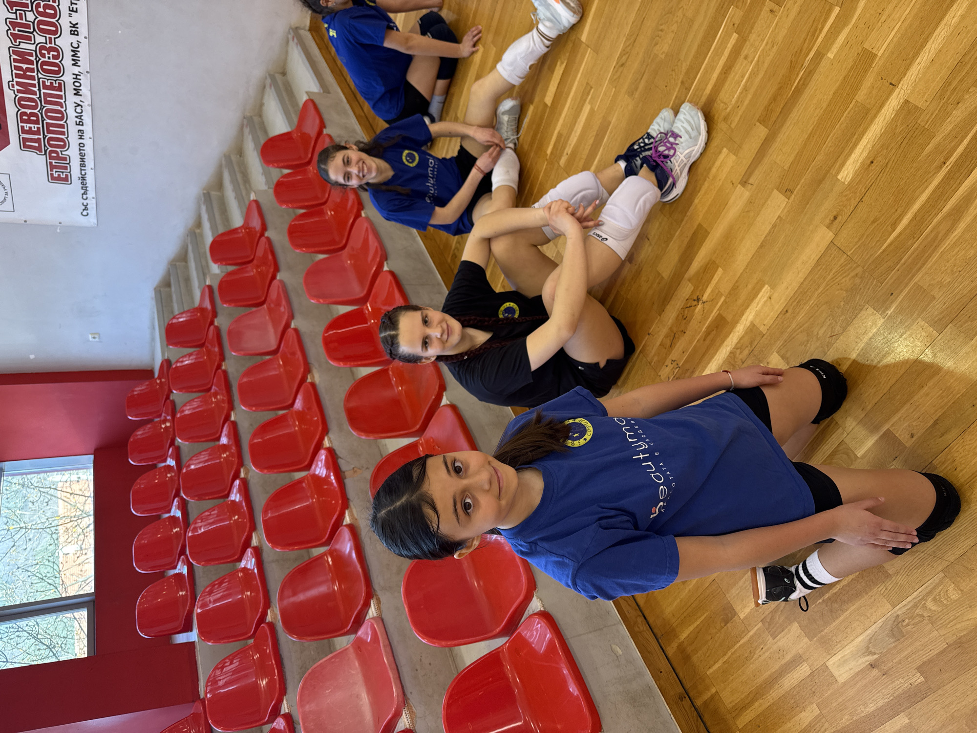 A group of four young female volleyball players are resting on the floor of a gymnasium in Etropole, Bulgaria. They are positioned near the bleacher seats. The players are wearing volleyball uniforms and knee pads. One player is wearing a blue T-shirt. A sign in the background identifies the location as the city of Etropole, Bulgaria and lists dates that include "ЕТРОПОЛЕ 03-06". There is a window to the left. The room is brightly lit.